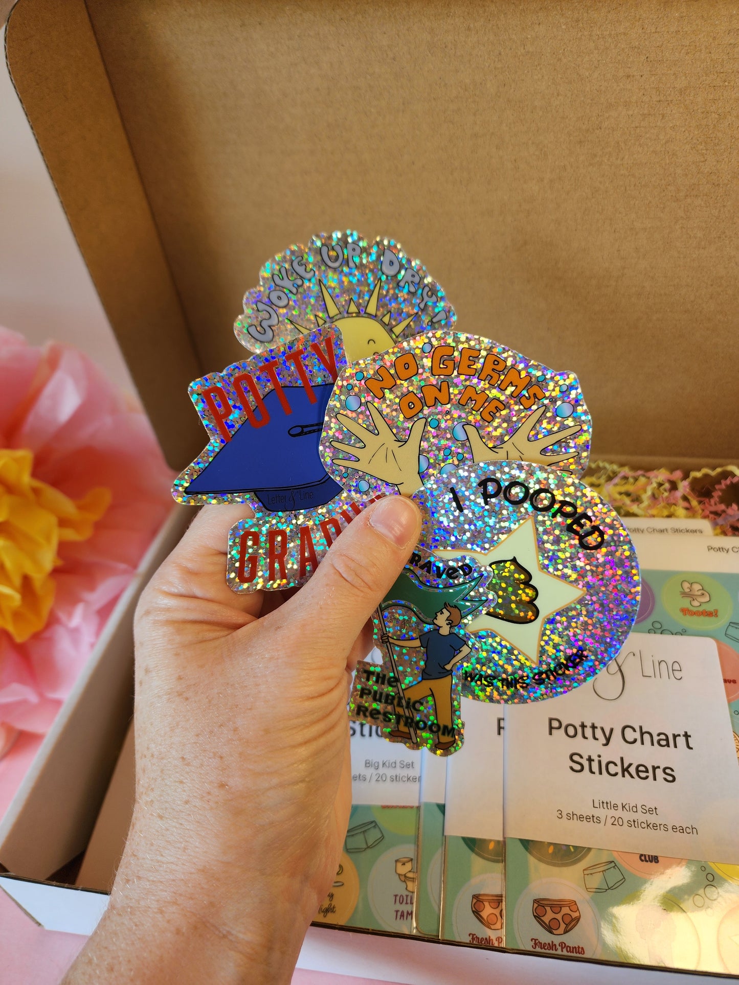 Hand holding all five included glitter stickers fanned out: Potty Graduate, Woke Up Dry, No Germs on Me, I Pooped, and I Braved the Public Restroom.