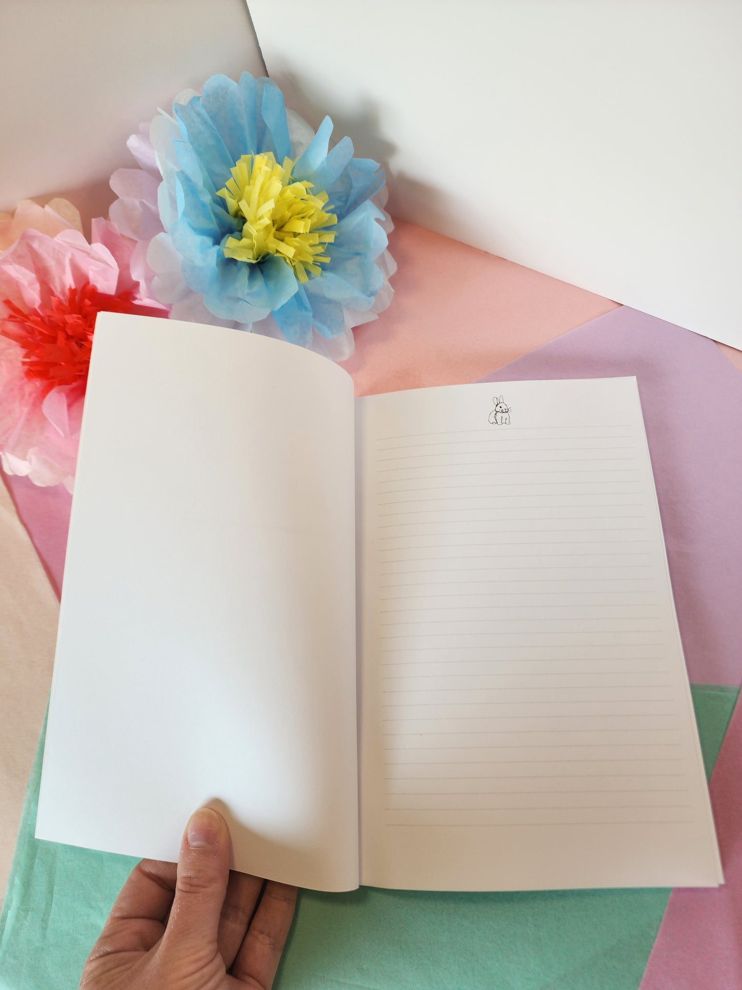 Interior first page of bunny notebook with lightly lined pages and a bunny illustration at the top of the page, held by a hand, with colorful flowers in the background.