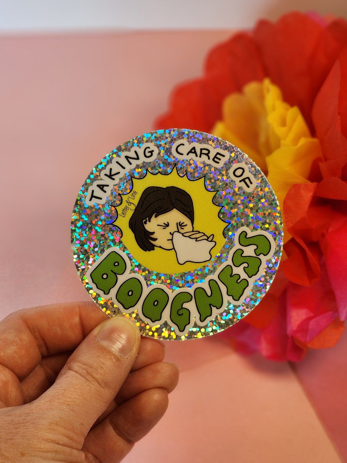 Hand holding a single “Taking Care of Boogness” round sticker in front of red, yellow, and pink tissue paper flowers.