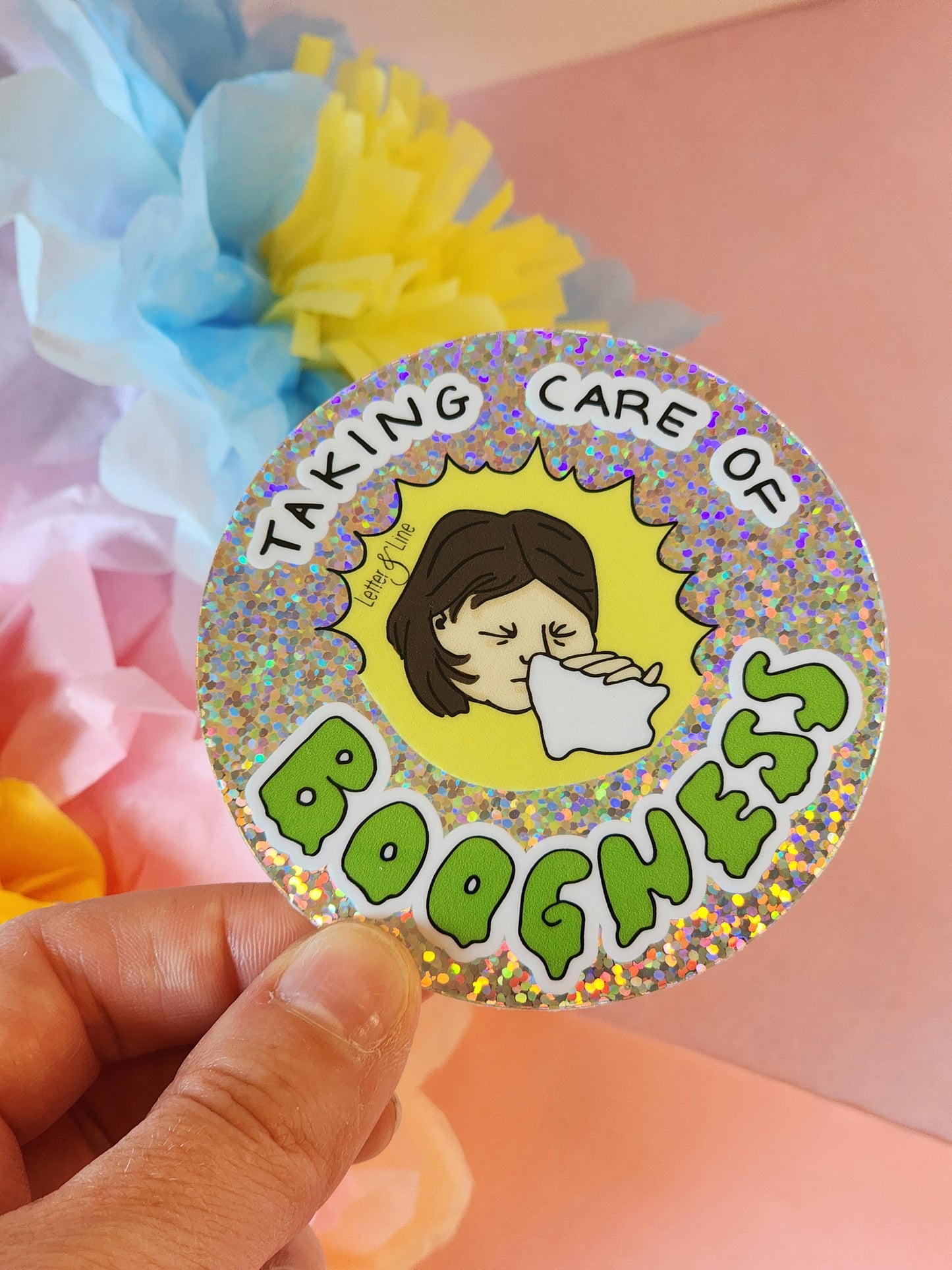 Hand holding a glittery “Taking Care of Boogness” sticker in front of pastel paper flowers on a pink background.