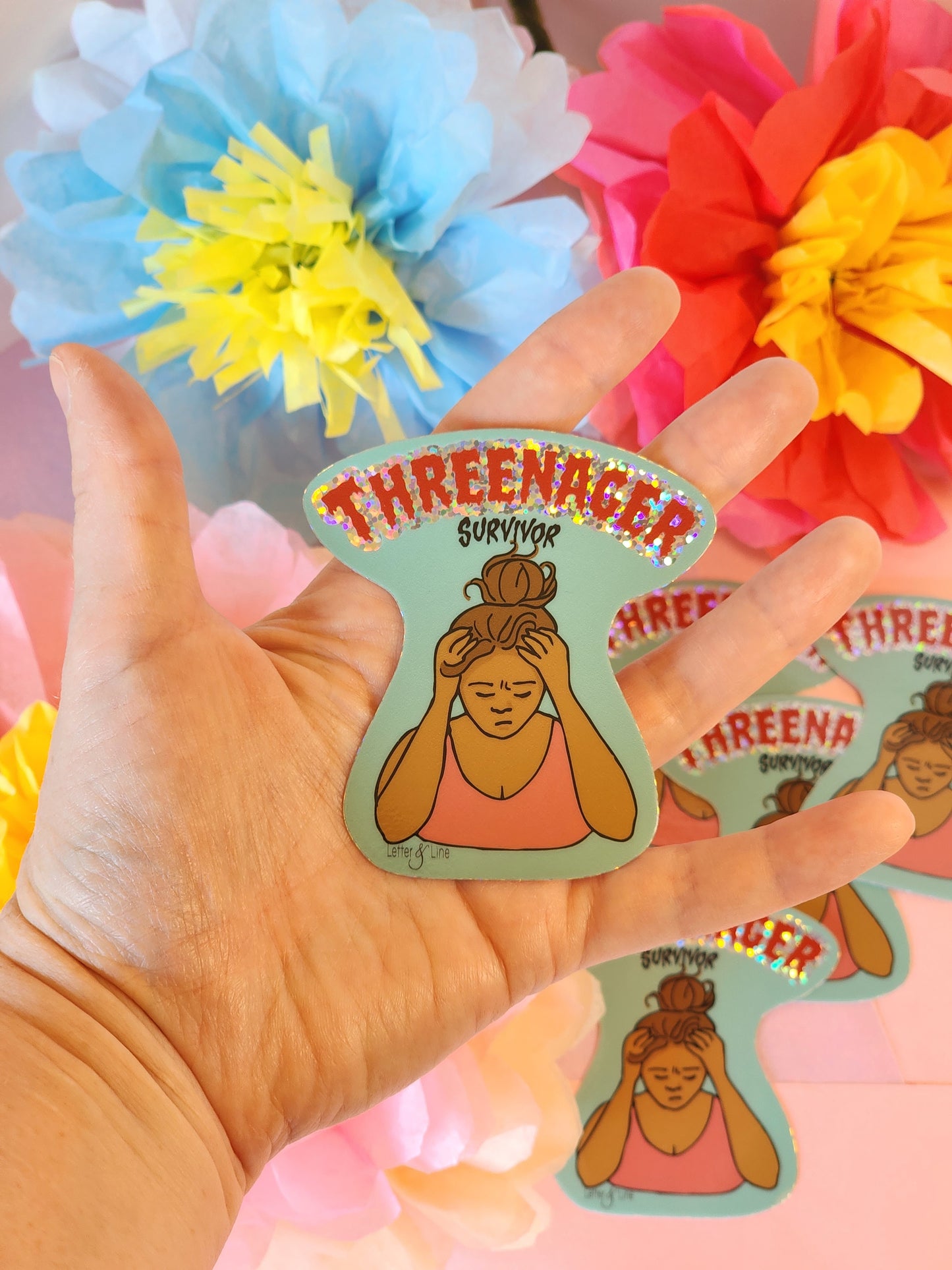 Hand holding the “Threenager Survivor” glitter sticker in front of vibrant tissue paper flowers in red, yellow, blue, and pink.