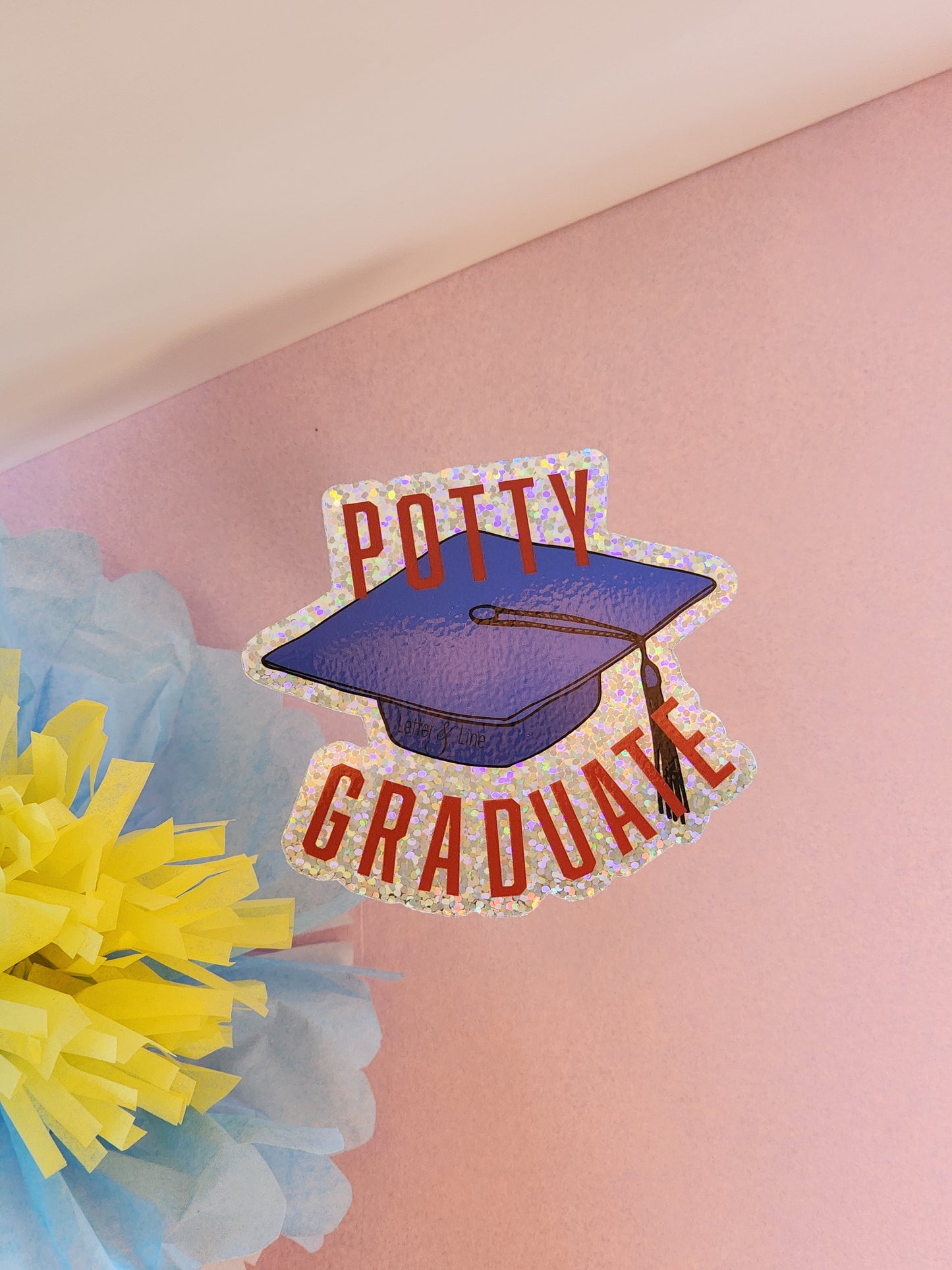 “Potty Graduate” glitter sticker with a graduation cap and red lettering placed on a pink surface, with pastel blue and yellow tissue flowers.