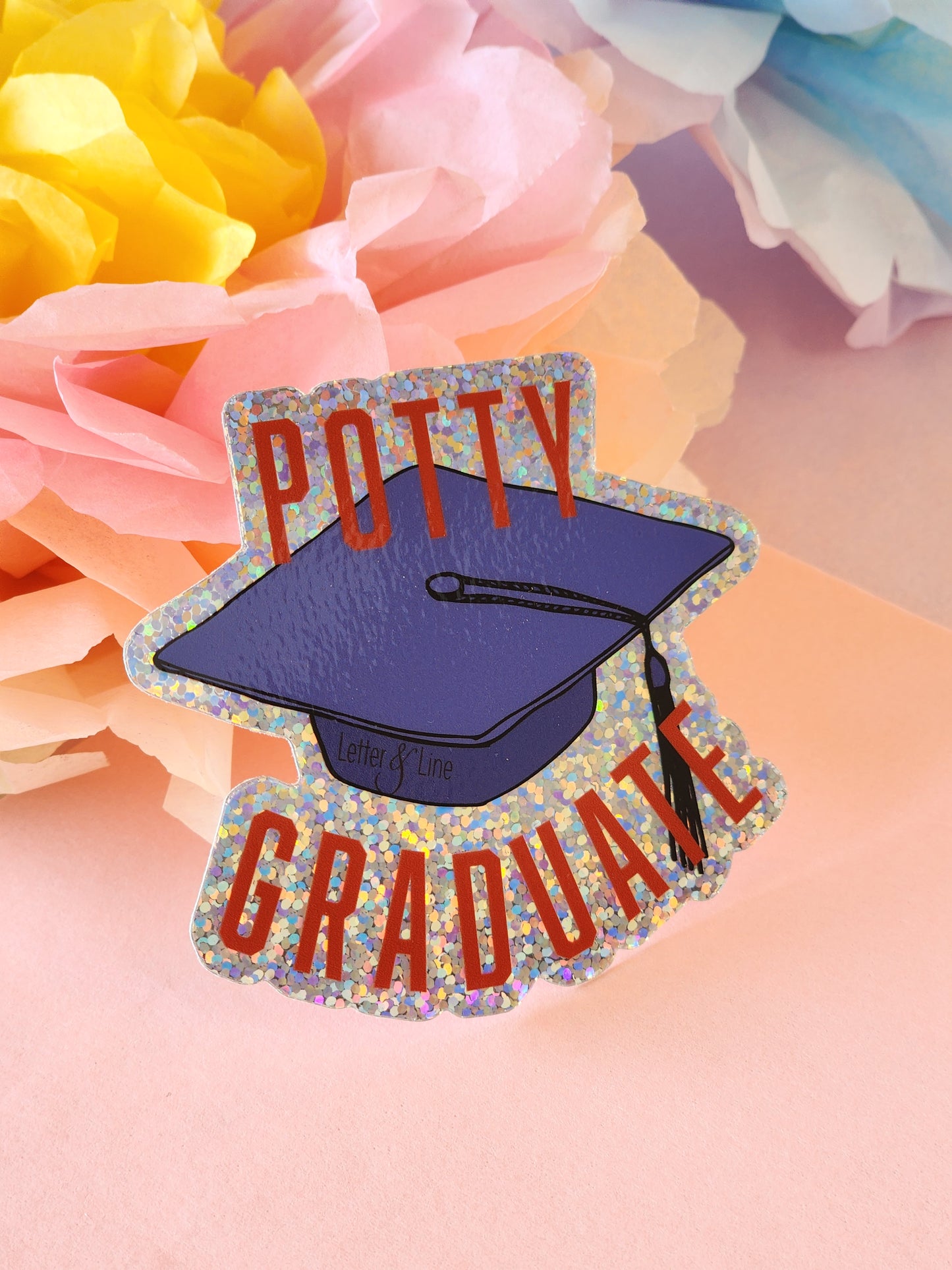 Close-up of a graduation cap “Potty Graduate” glitter sticker on a pink background with peach, yellow, and blue paper flowers in the corners.