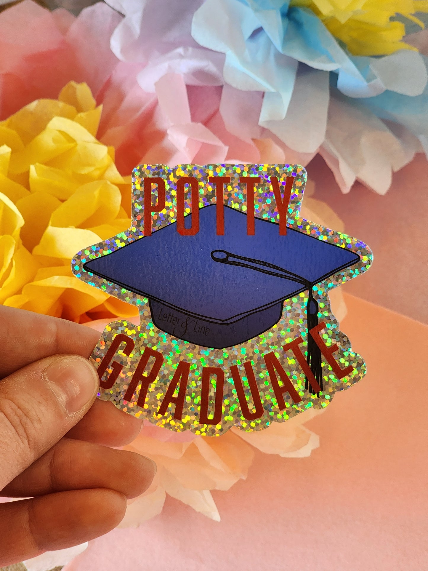 Hand holding a “Potty Graduate” glitter sticker with a bold graduation cap design, in front of yellow and blue tissue flowers.