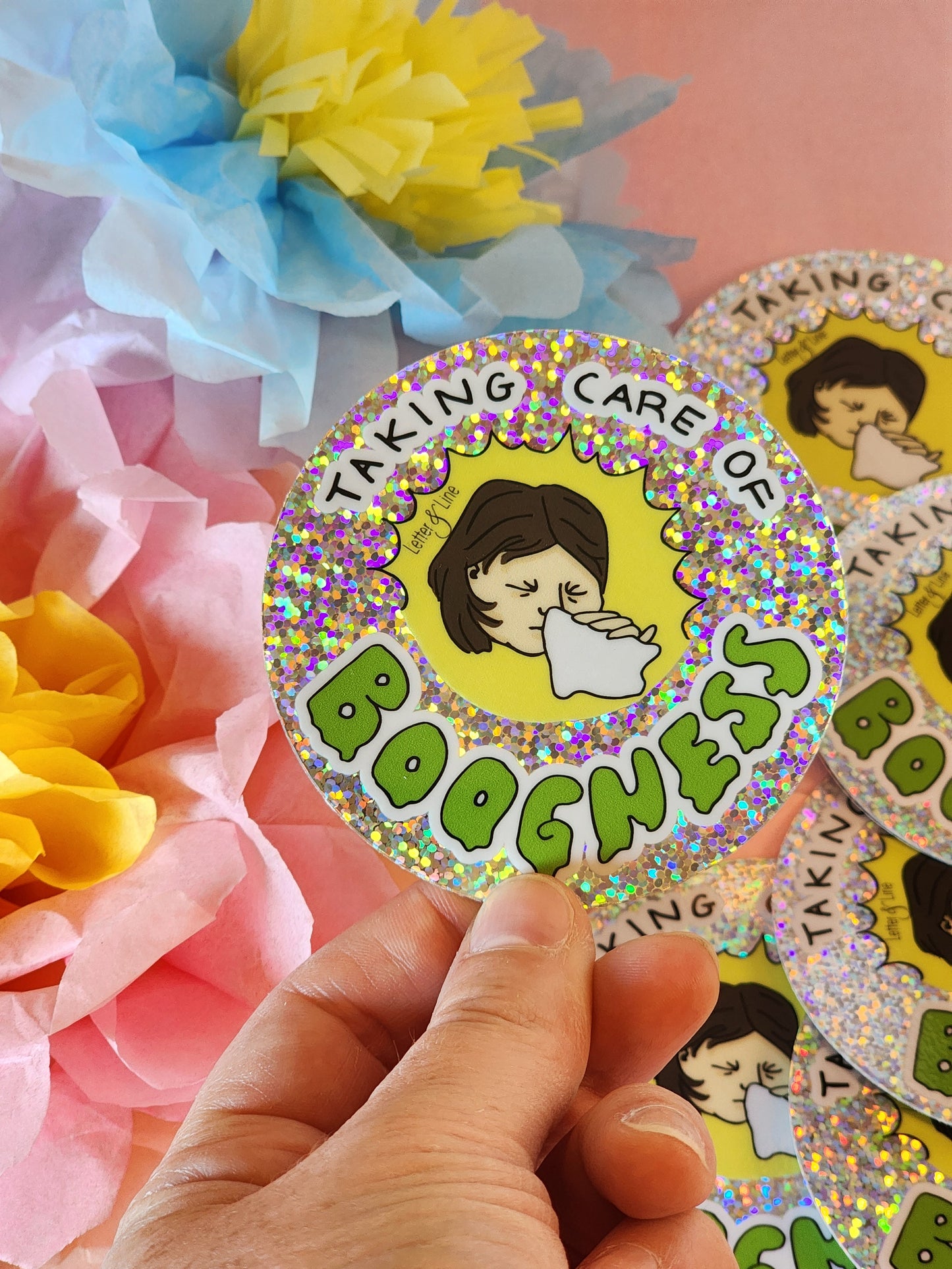 Hand holding a “Taking Care of Boogness” glitter sticker over a group of matching stickers, with pastel flowers in the background.