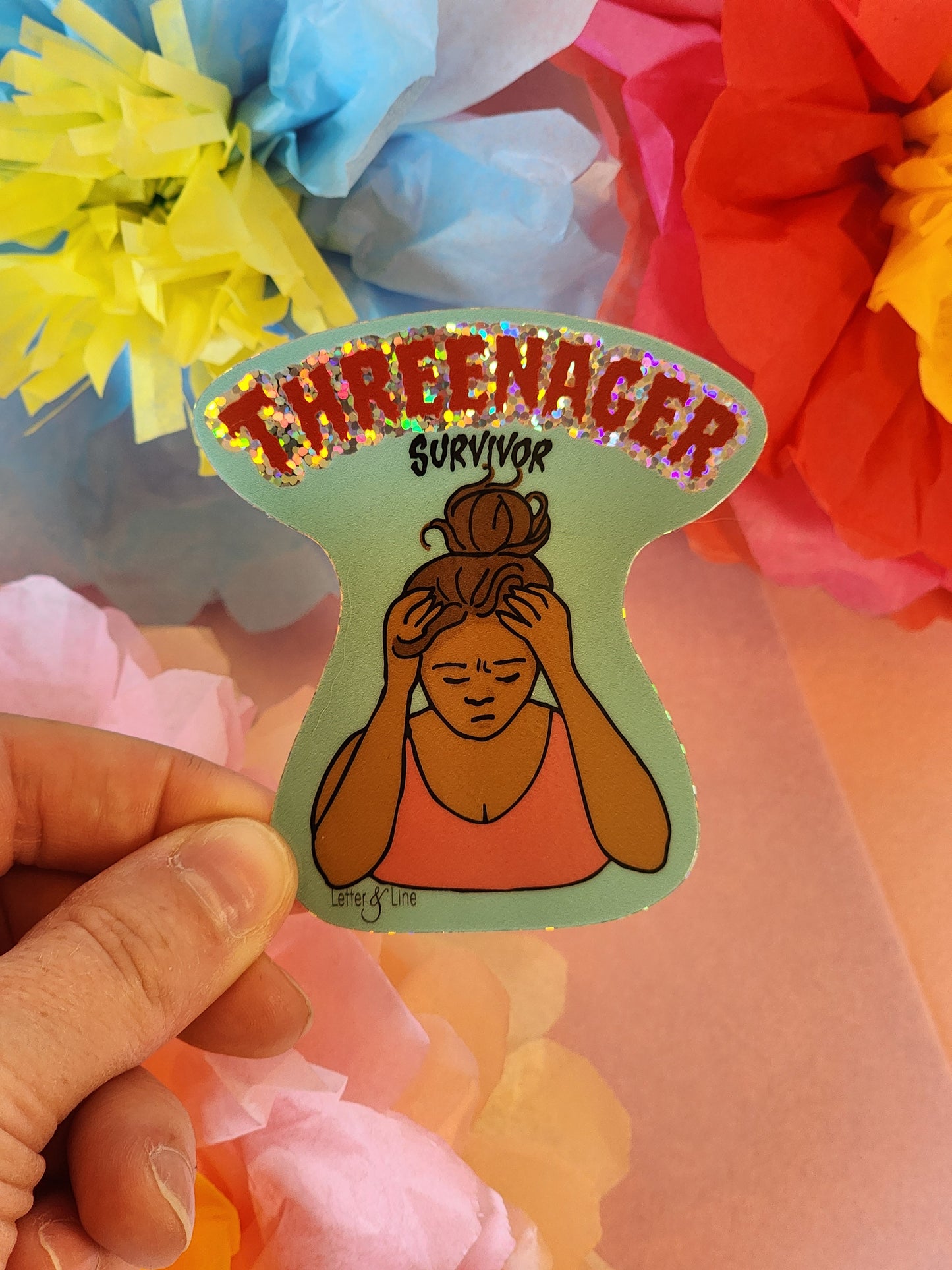 Close-up of stressed out mom “Threenager Survivor” glitter sticker held in front of red and yellow tissue flowers on a pink backdrop.