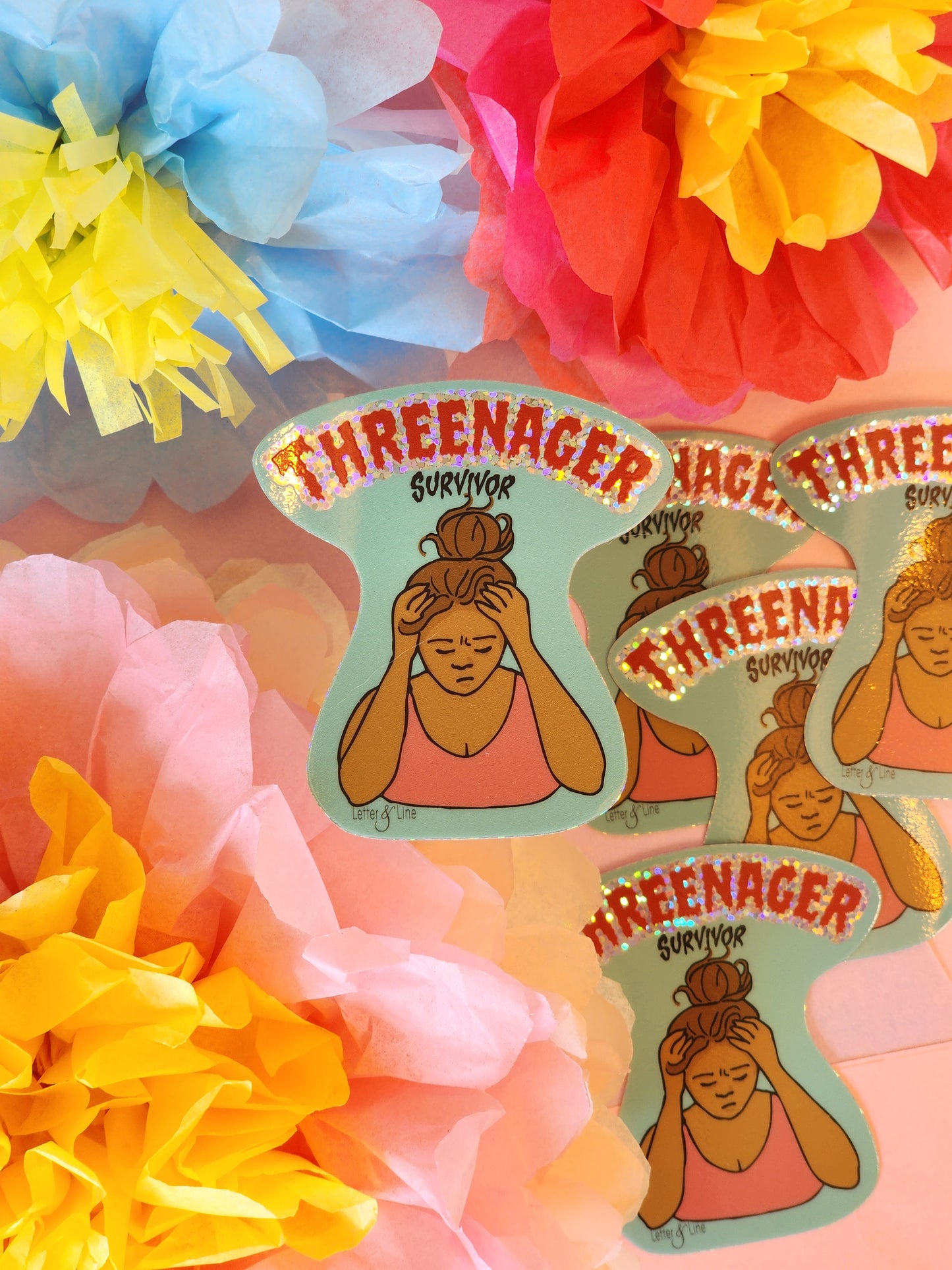 Stressed out mom illustration holding head in hands with text “Threenager Survivor” on a glitter sticker. Duplicate stickers arranged in the background on a pink surface surrounded by layered tissue flowers in yellow, pink, and blue.