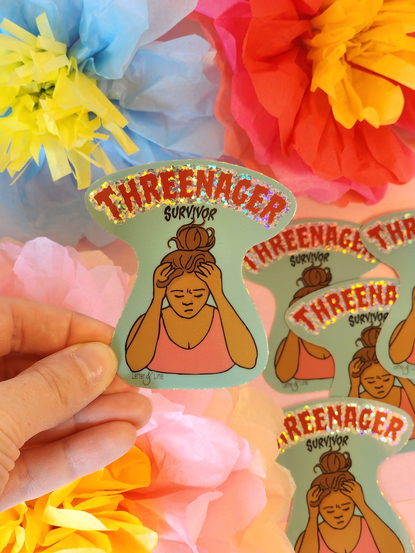 Hand holding a “Threenager Survivor” glitter sticker with a messy exasperated mom illustration with multiple matching stickers in background.