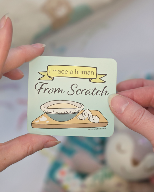 Hand holding I Made a Human From Scratch new parent milestone sticker showing illustrated mixing bowl and banner design