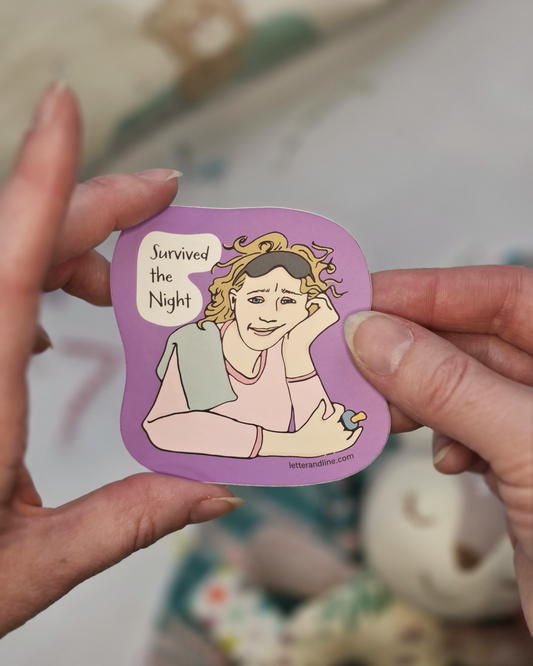 Hand holding Survived the Night new parent milestone sticker showing illustrated exhausted mom design on purple background