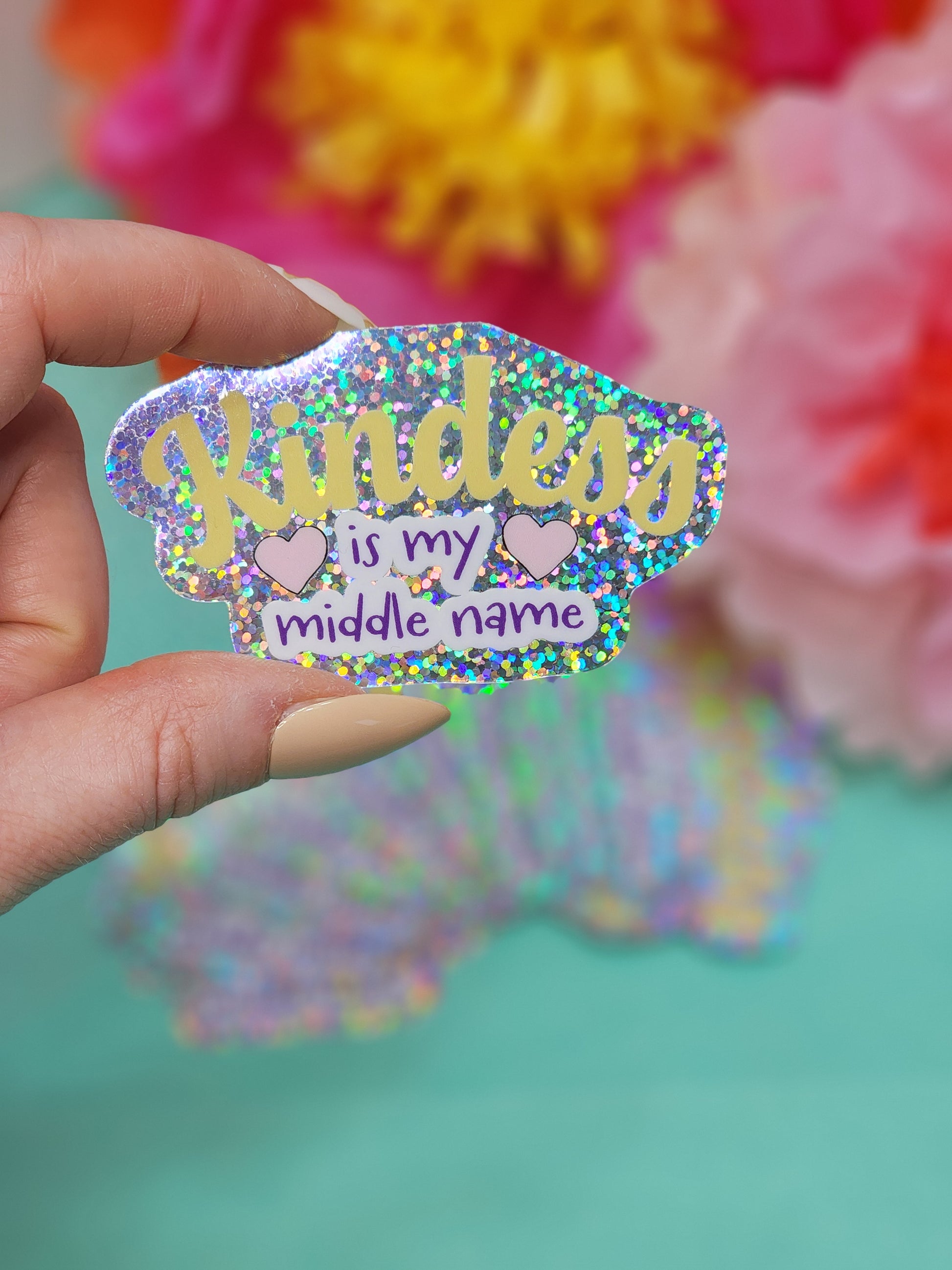 hand holding a single “Kindness is my middle name” glitter sticker in front of pink and yellow flowers
