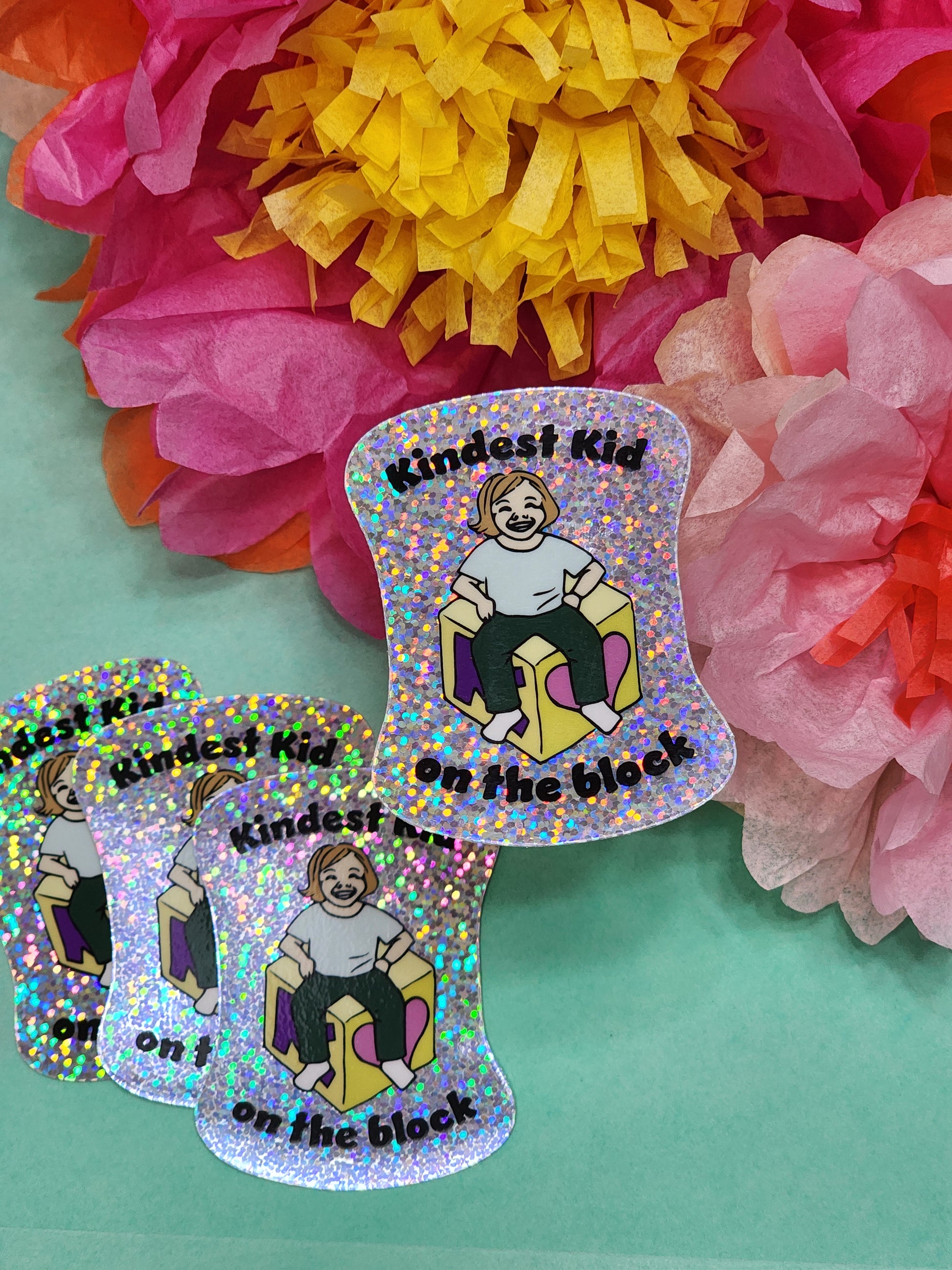 group of “Kindest Kid on the Block” glitter stickers arranged on teal with colorful tissue flowers