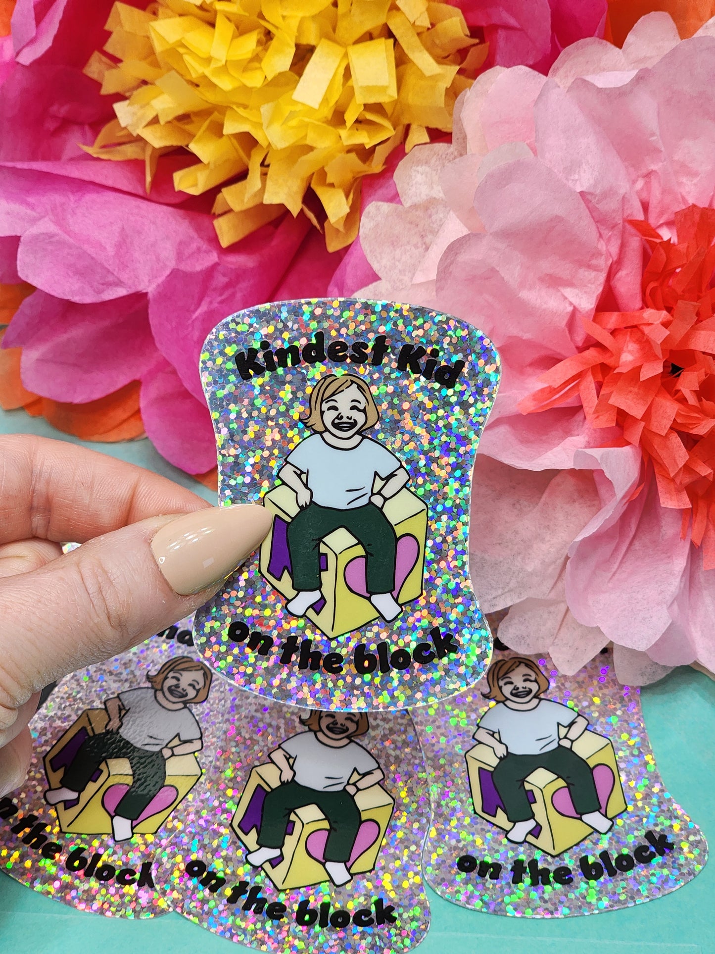 hand holding a “Kindest Kid on the Block” glitter sticker above a small stack of matching stickers
