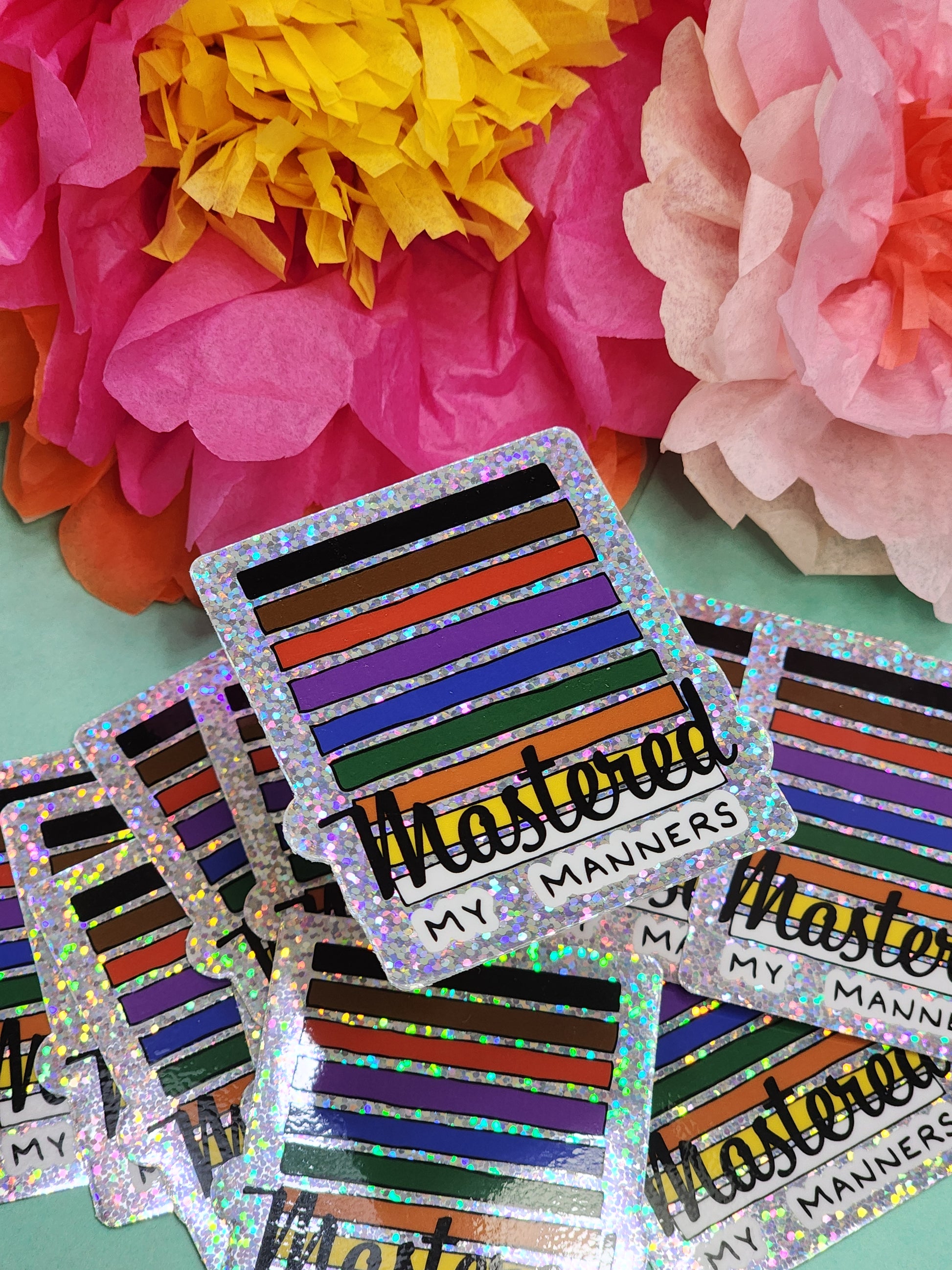 pile of “Mastered my Manners” glitter stickers arranged on teal with pink and yellow tissue flowers