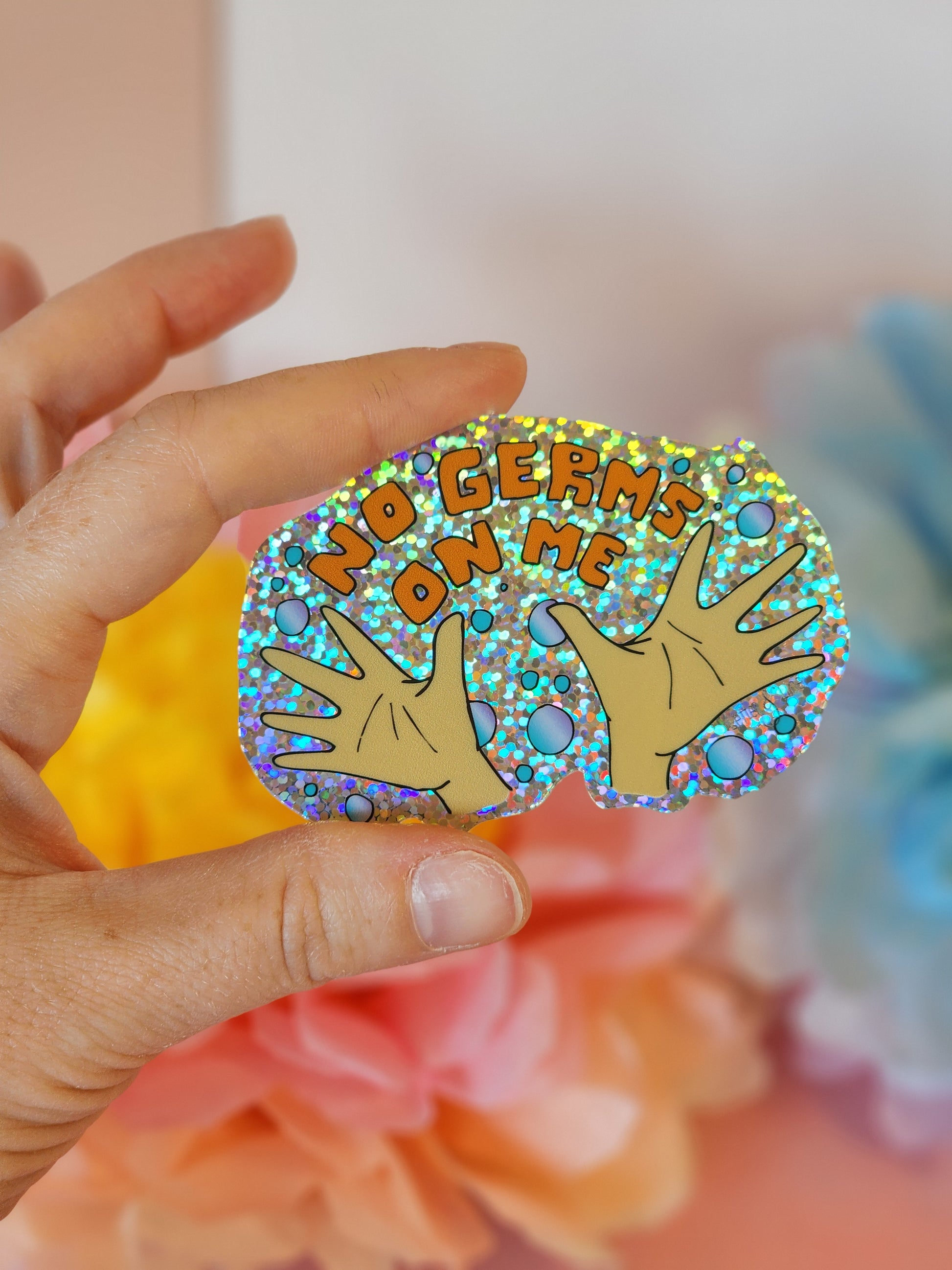 Hand holding a glittery “No Germs on Me” sticker with cartoon hands and bubbles, in front of pastel tissue flowers.