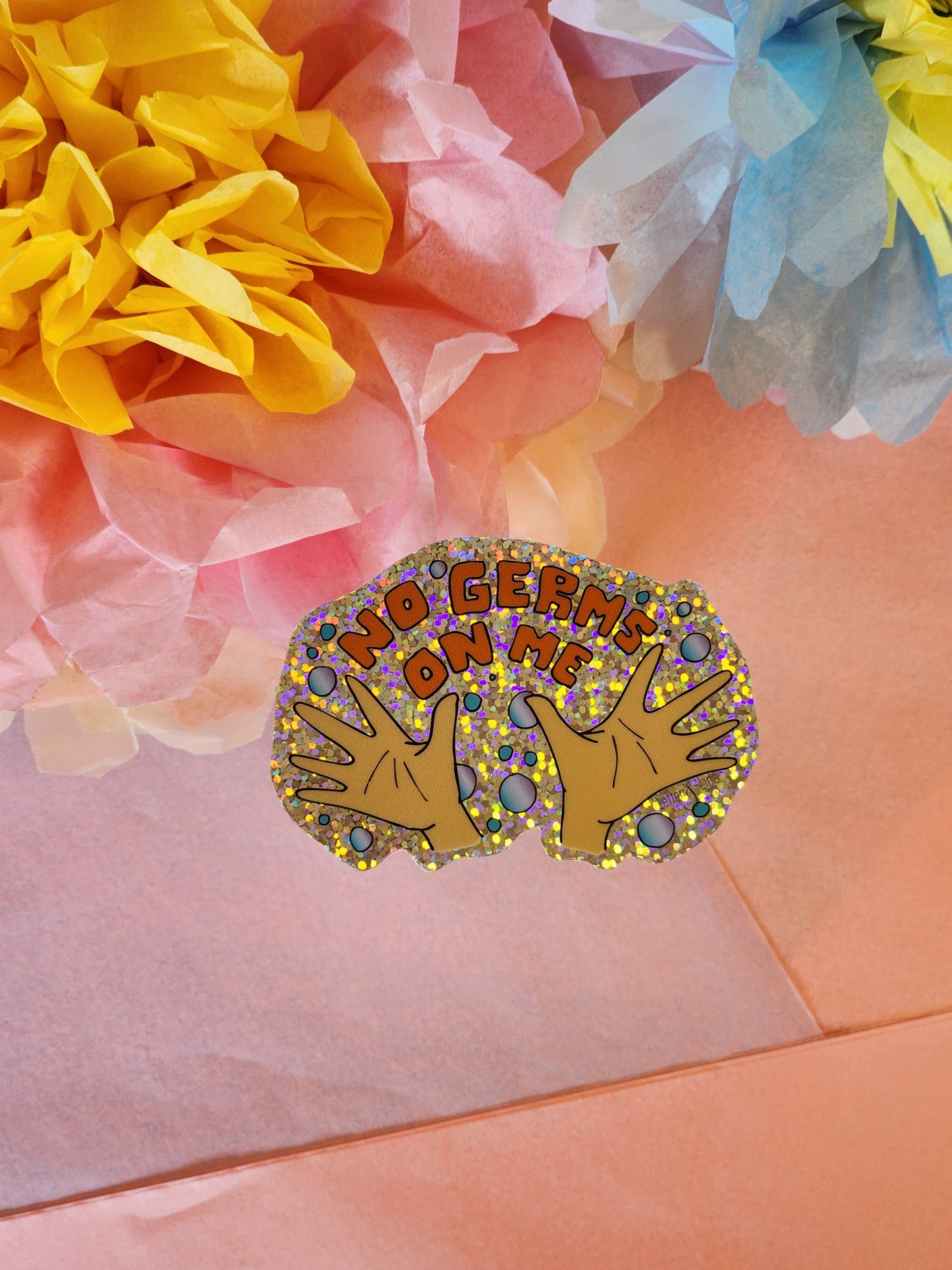 Single “No Germs on Me” glitter sticker showing clean hands and bubbles, placed on a pink surface in front of colorful tissue flowers.