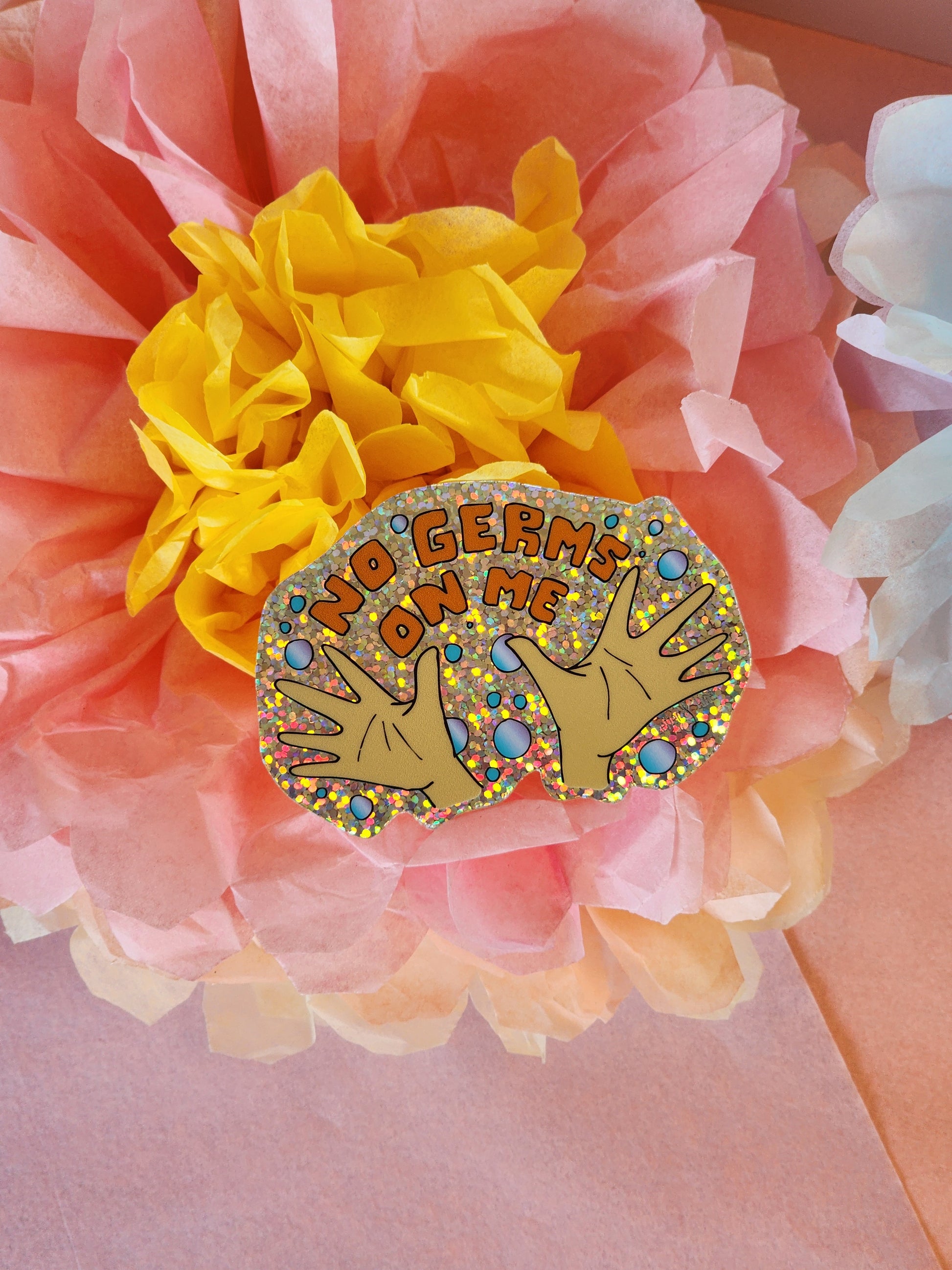 Single glittery “No Germs on Me” sticker showing hands and bubbles with orange text, lying on a yellow, pink, and white tissue paper flower. 