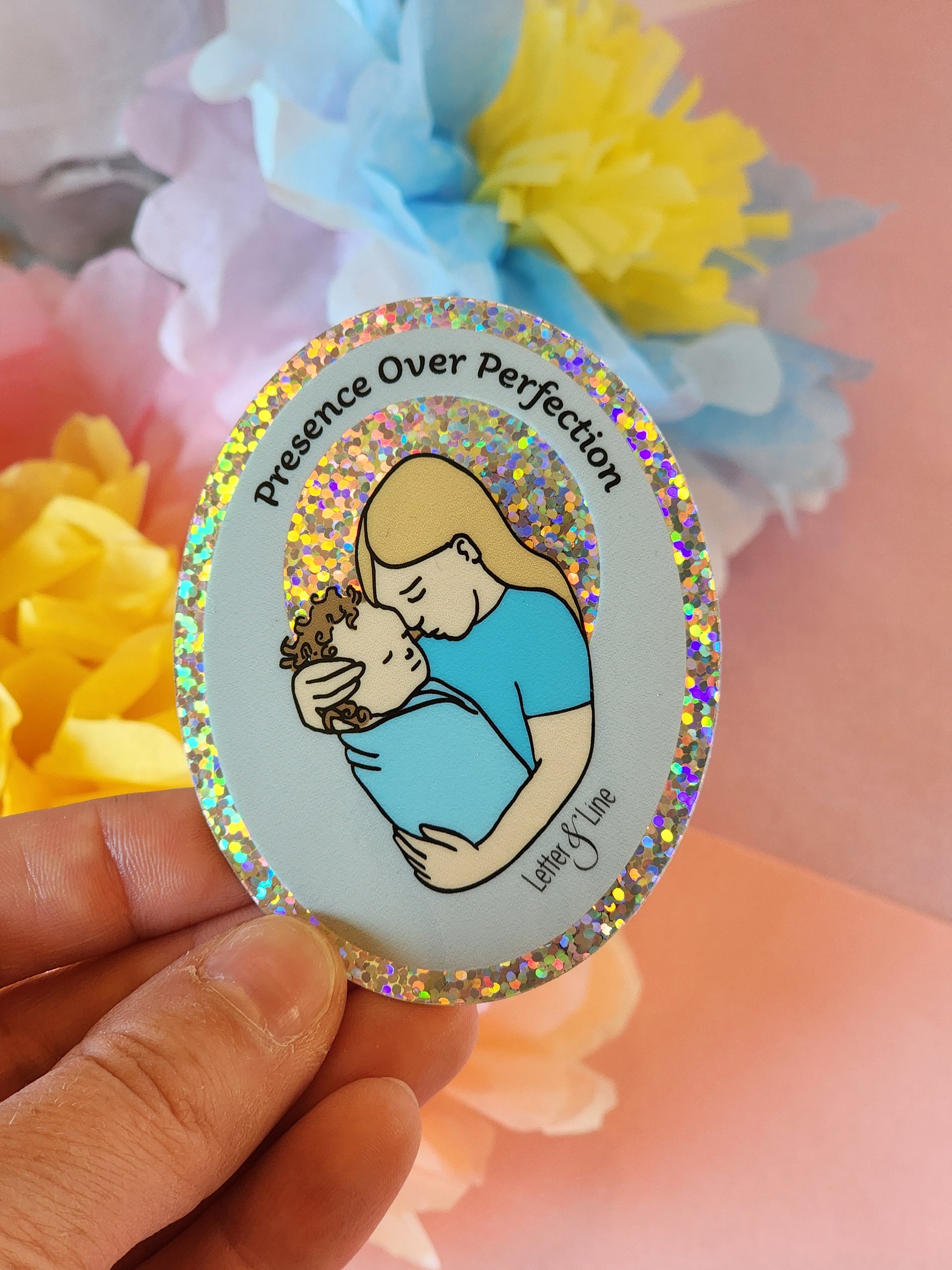 Hand holding a glittery “Presence Over Perfection” sticker featuring a mother cradling her child, with pastel tissue flowers in the background.