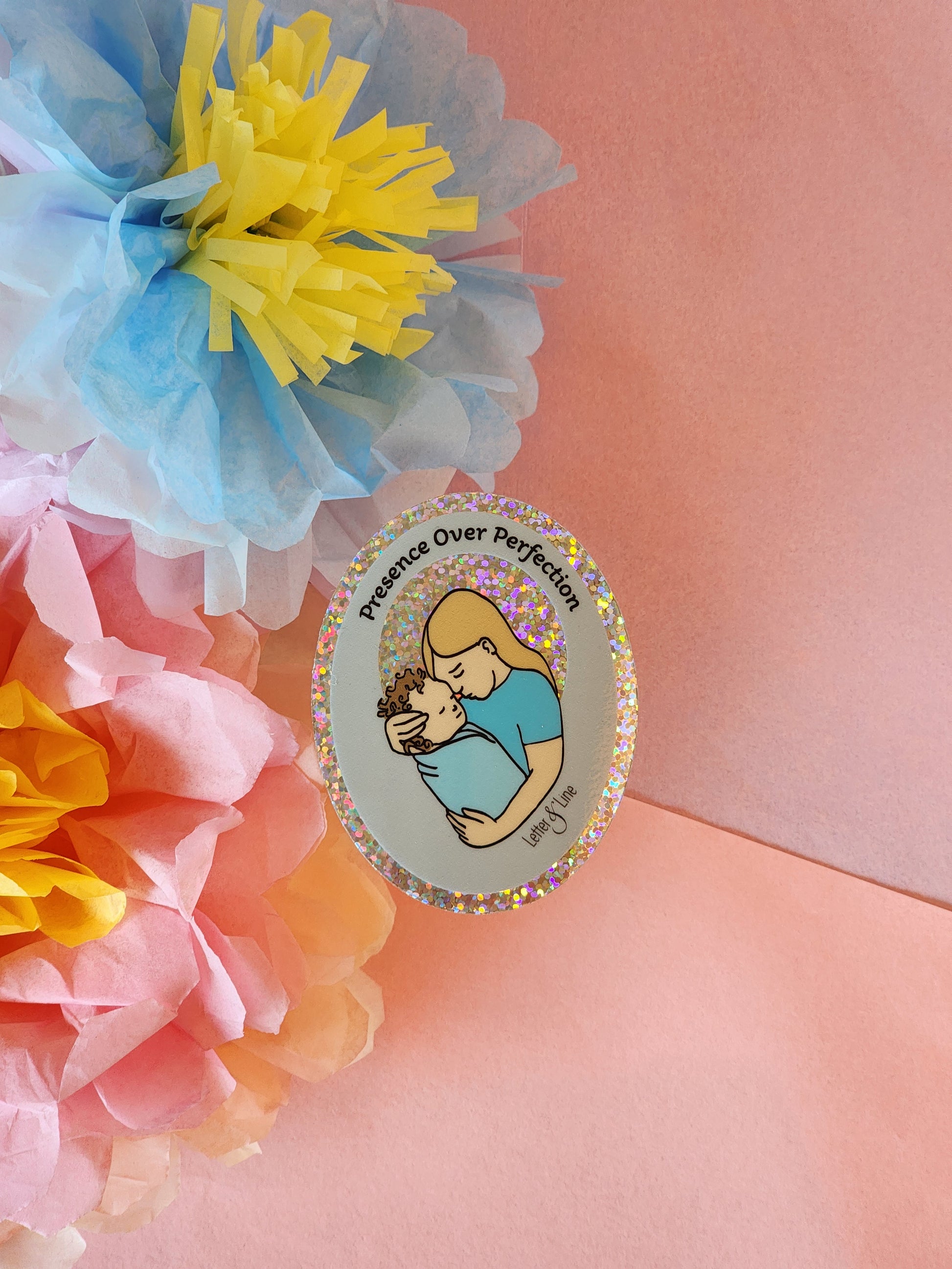 One “Presence Over Perfection” mother and baby sticker placed on a soft pink surface, surrounded by blue, yellow, and pink paper flowers.