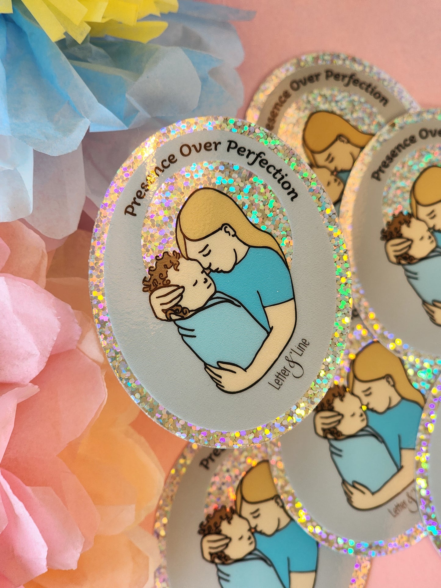 Several “Presence Over Perfection” glitter stickers, featuring a mother hugging her infant, layered in a group, with pastel flowers on a pink background.