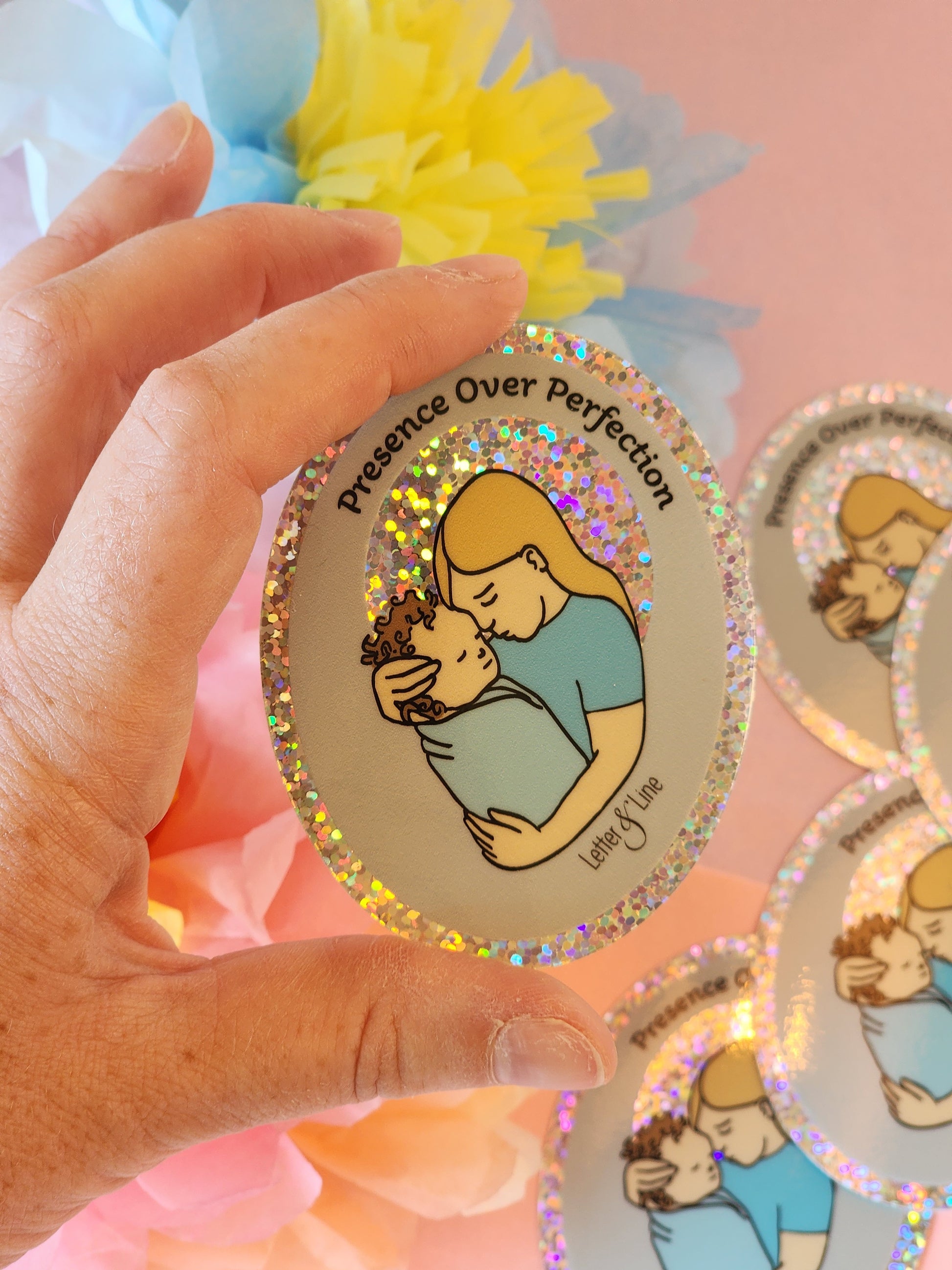 Hand holding a “Presence Over Perfection” sticker above a pile of matching stickers, with soft floral decor behind. Sticker features a blonde woman holding a curly-haired brunette baby.