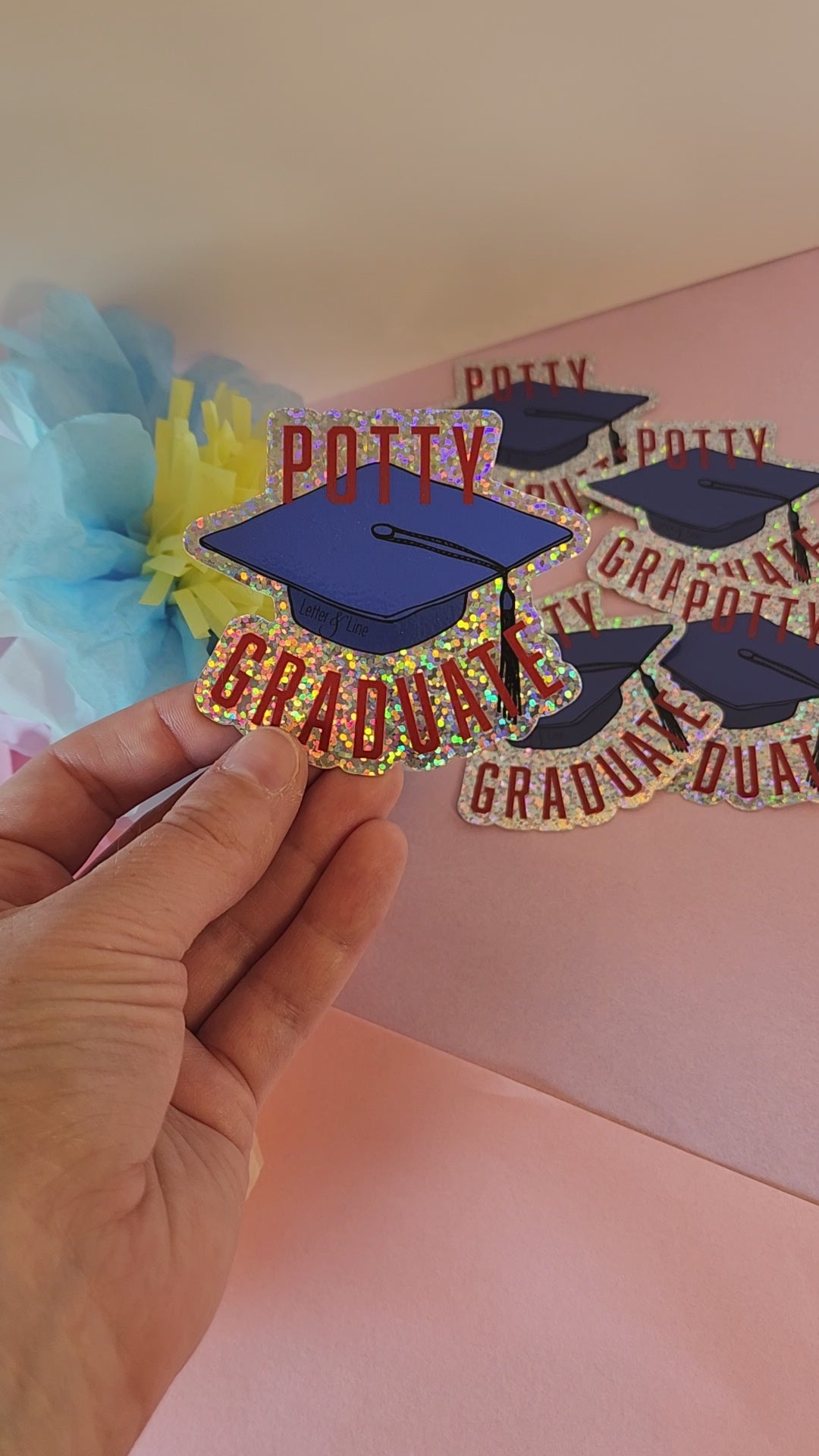 Video preview of a hand turning the “Potty Graduate” sticker to show its glitter effect, set against a soft floral background.