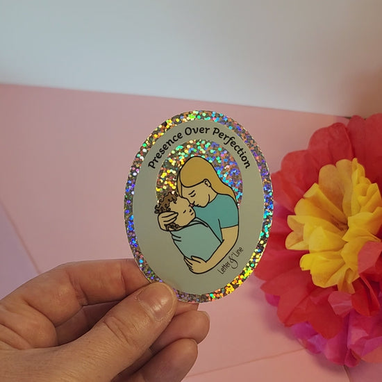 Video of a hand rotating the “Presence Over Perfection” sticker to show its glittery vinyl shine, set against pastel paper flowers.