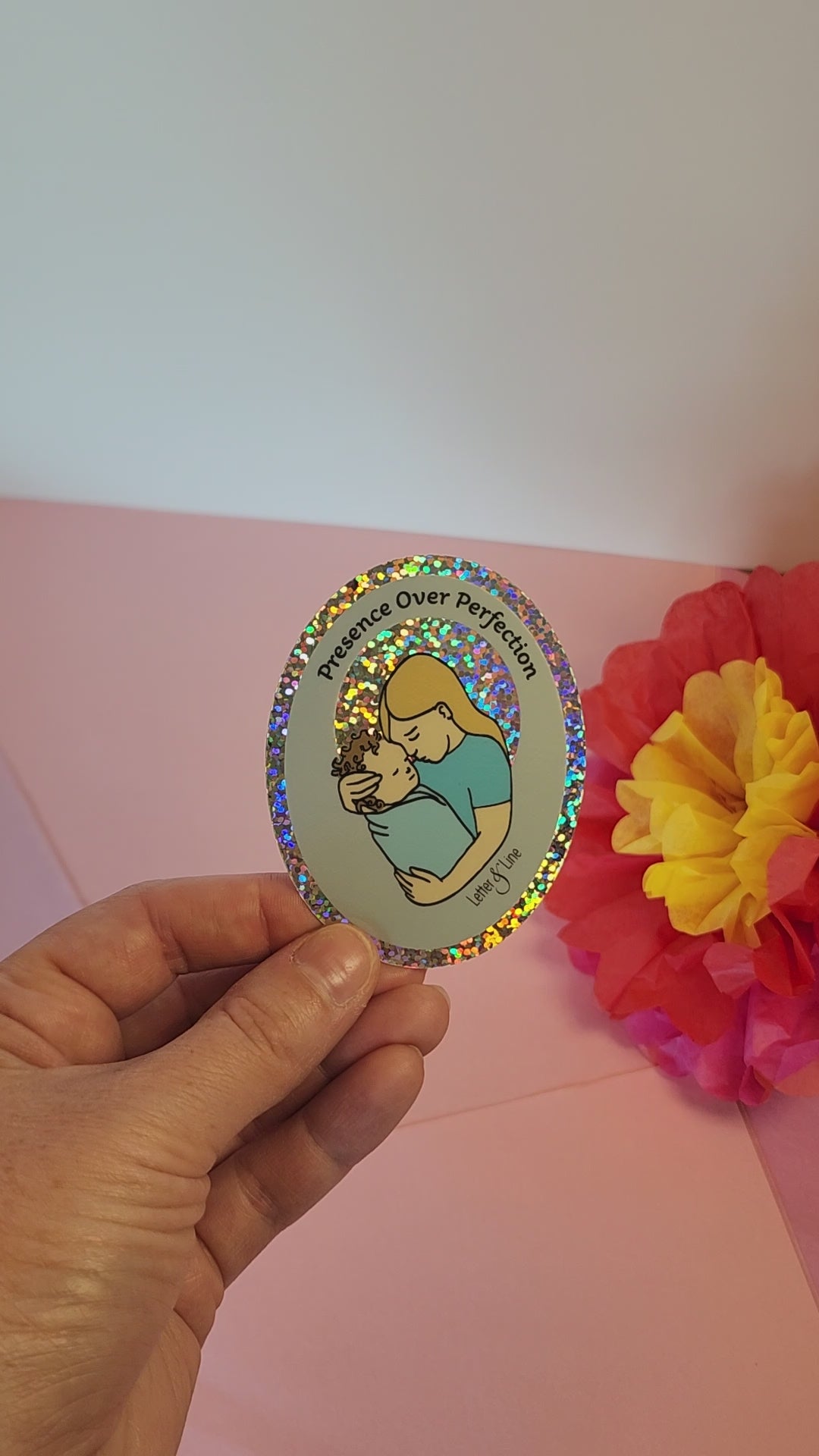 Video of a hand rotating the “Presence Over Perfection” sticker to show its glittery vinyl shine, set against pastel paper flowers.