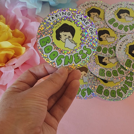 Video showing a hand rotating the “Taking Care of Boogness” sticker to highlight the glitter finish, with colorful flowers behind.