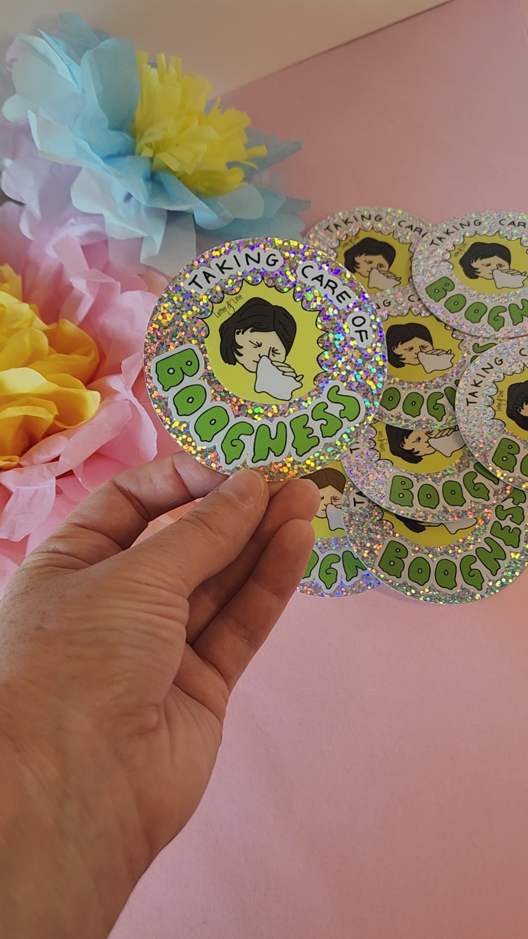 Video showing a hand rotating the “Taking Care of Boogness” sticker to highlight the glitter finish, with colorful flowers behind.