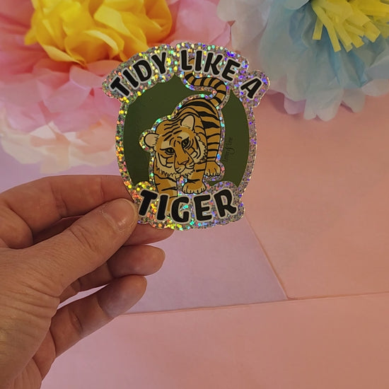 Video showing hand rotating the “Tidy Like a Tiger” sticker to reveal its glitter sparkle, with flower decor in background.
