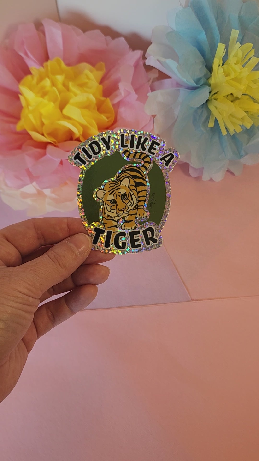 Video showing hand rotating the “Tidy Like a Tiger” sticker to reveal its glitter sparkle, with flower decor in background.