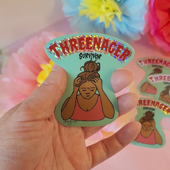 Video of a hand turning the “Threenager Survivor” sticker to catch the glitter shine, with soft floral background.