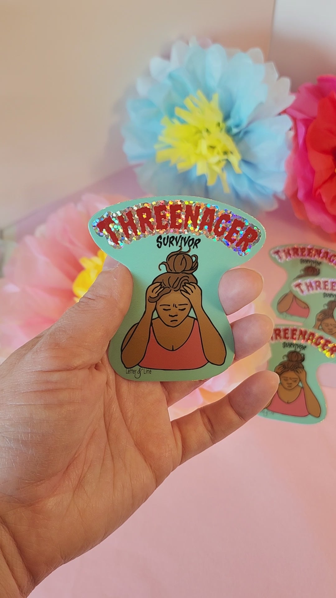 Video of a hand turning the “Threenager Survivor” sticker to catch the glitter shine, with soft floral background.
