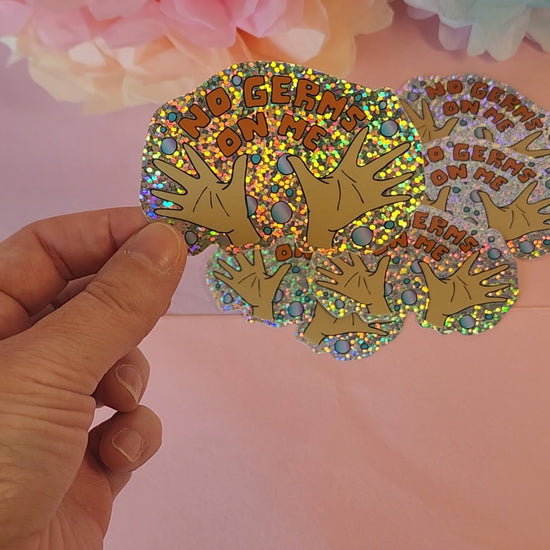 Video of a hand rotating the “No Germs on Me” sticker to show its glittery shine, with pastel floral background.
