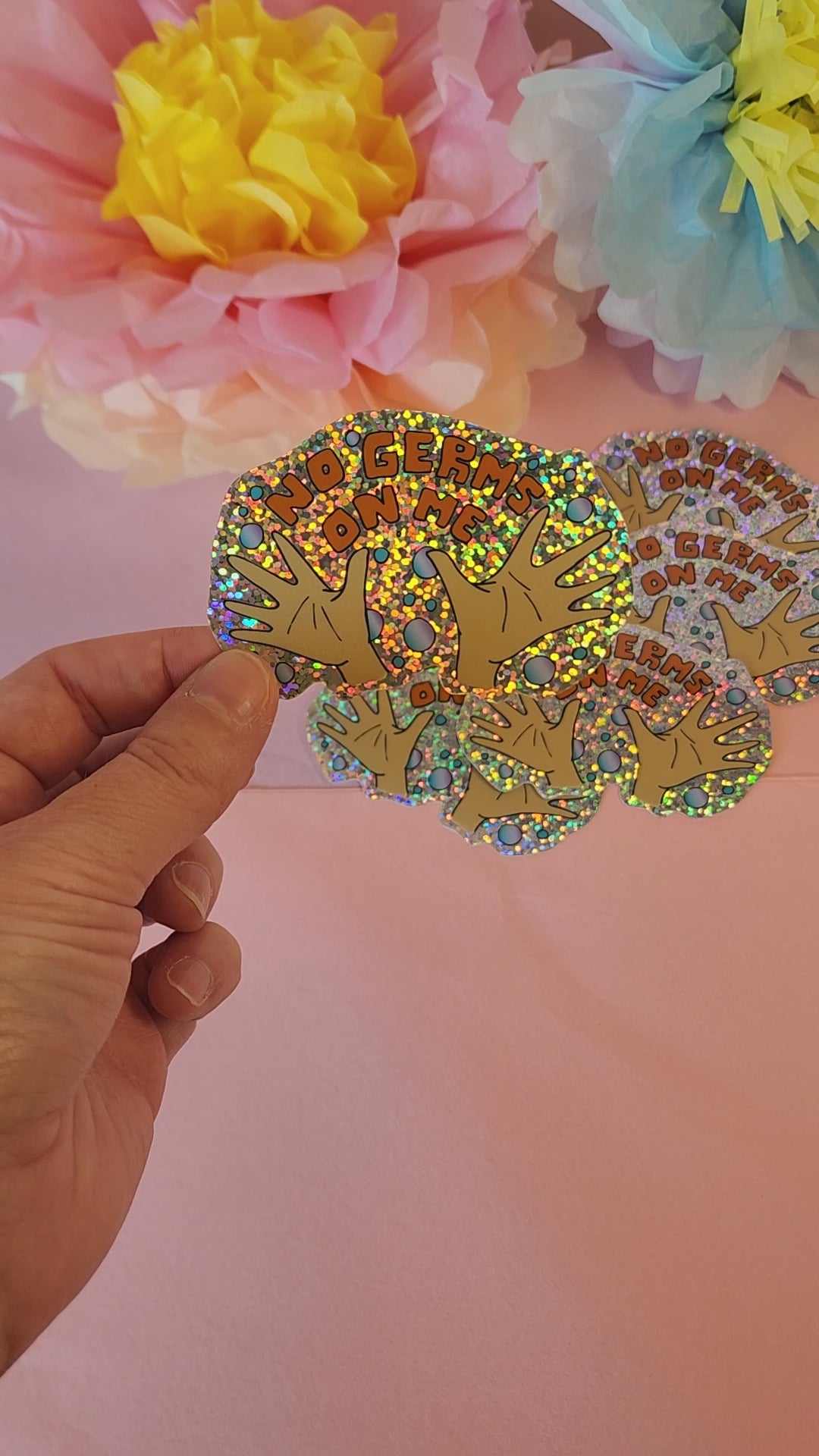 Video of a hand rotating the “No Germs on Me” sticker to show its glittery shine, with pastel floral background.