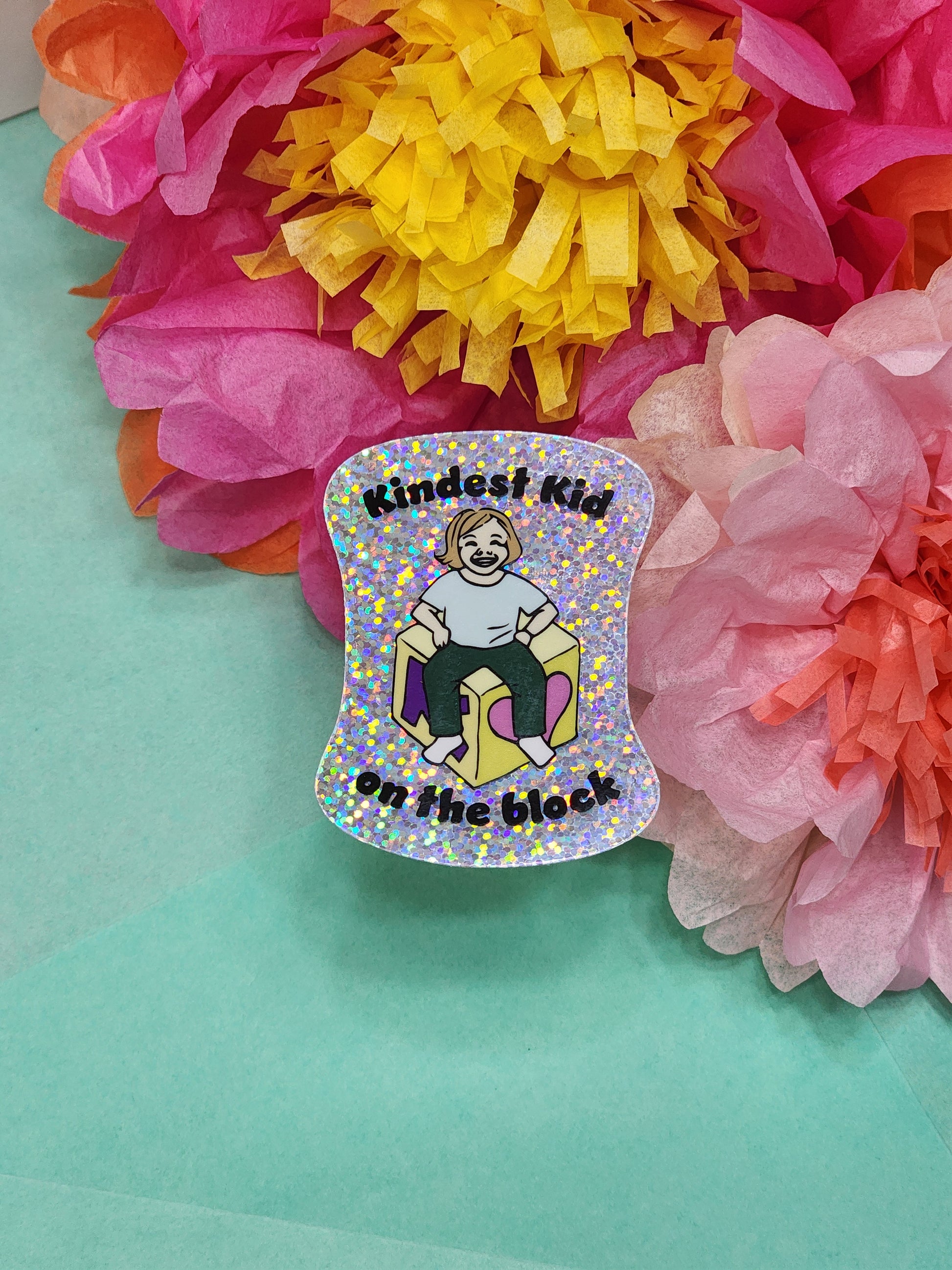 glitter “Kindest Kid on the Block” sticker on teal background with bright tissue flowers