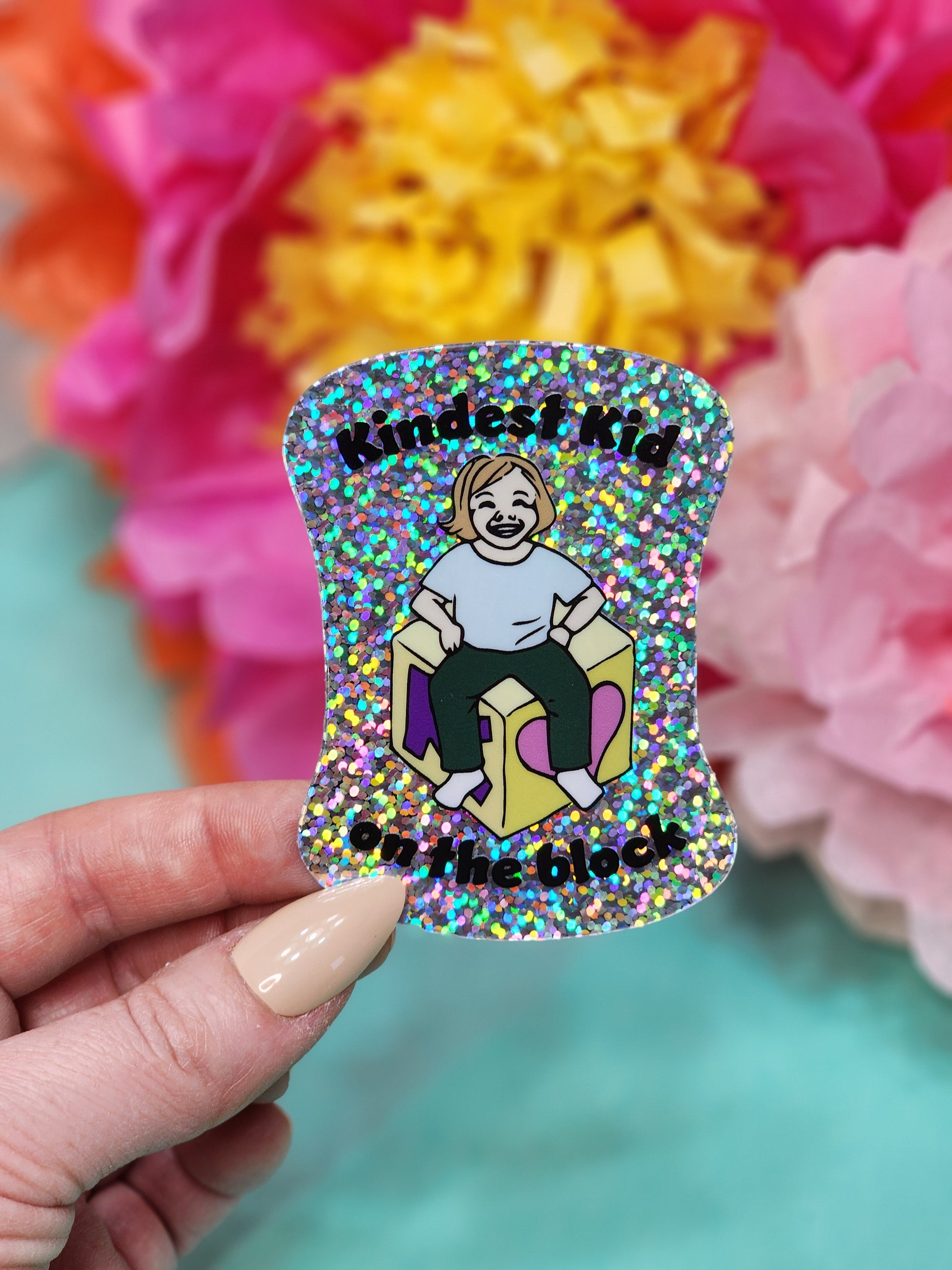 hand holding a single “Kindest Kid on the Block” glitter sticker in front of pink and orange flowers