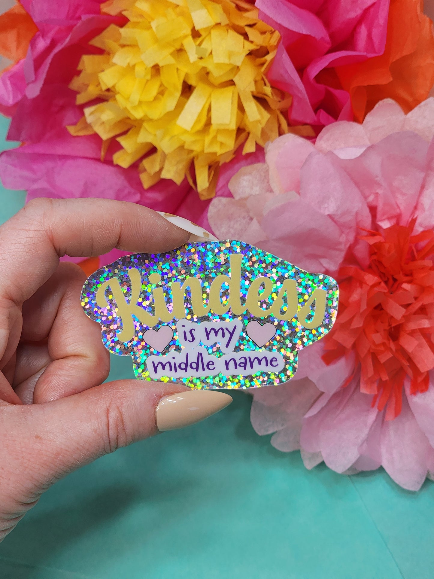 glitter “Kindness is my middle name” sticker held in hand with bright tissue flowers on teal background