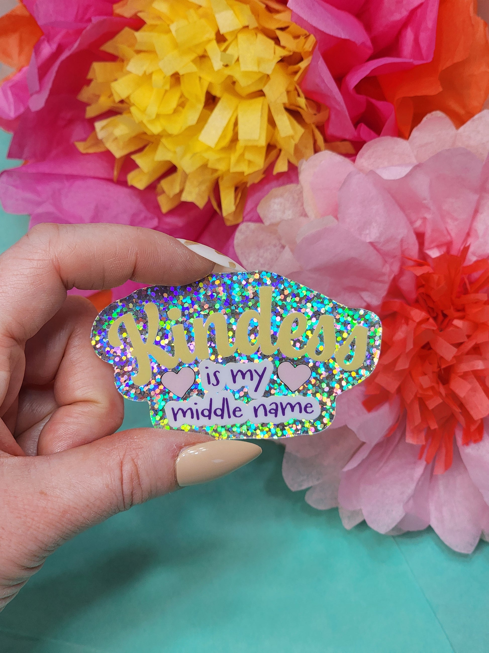 glitter “Kindness is my middle name” sticker held in hand with bright tissue flowers on teal background
