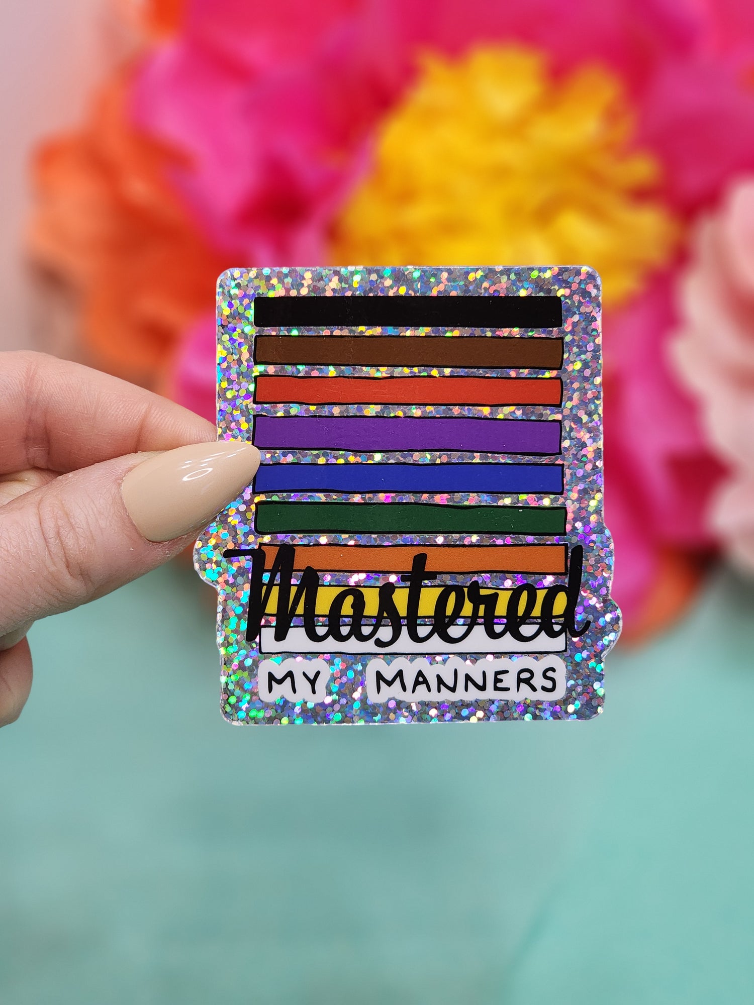 hand holding a glitter “Mastered my Manners” sticker with rainbow belt-style stripes on teal background with tissue flowers