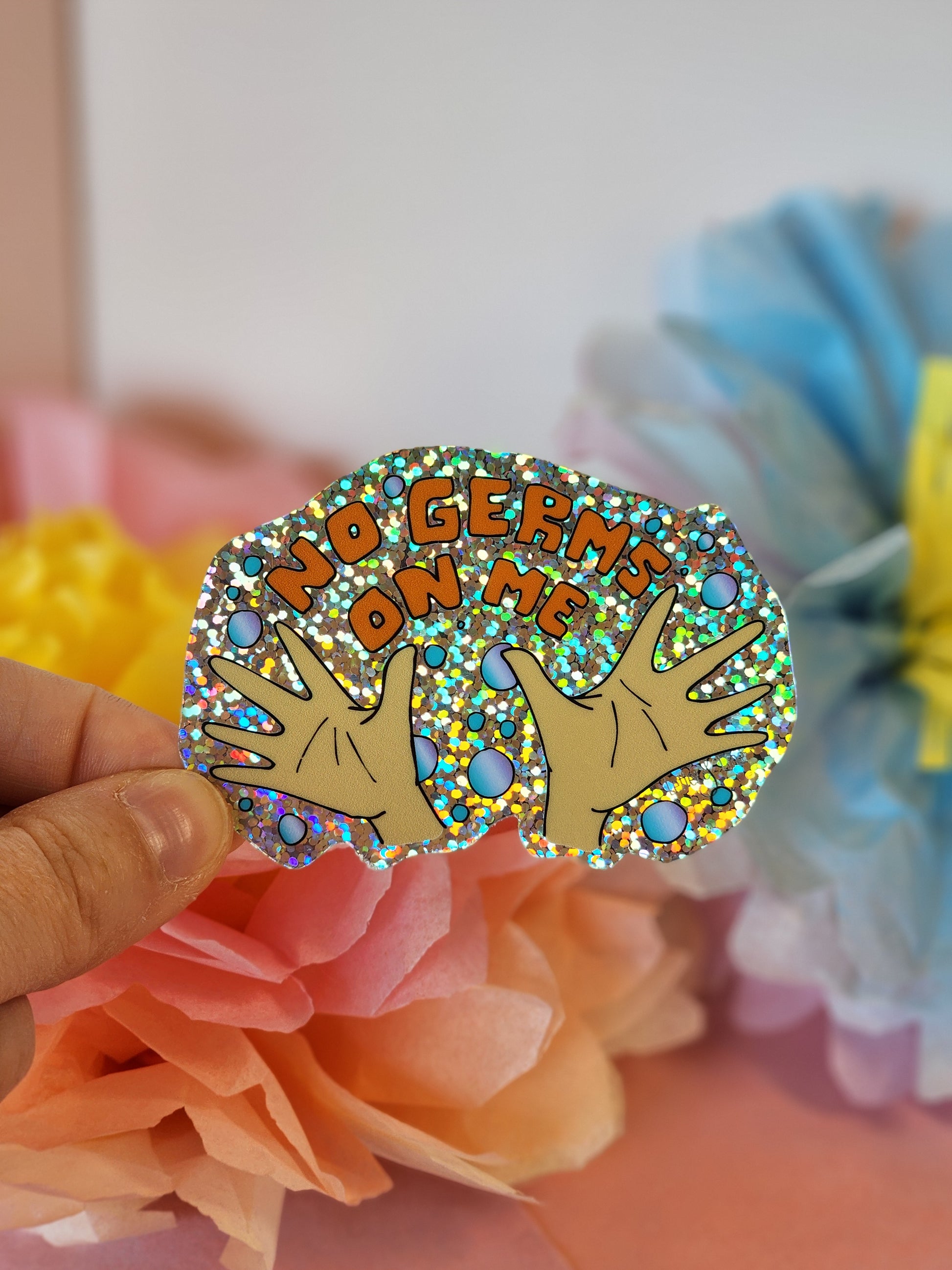 Hand holding the “No Germs on Me” glitter sticker in front of pastel blue and yellow paper flowers on a soft pink background.