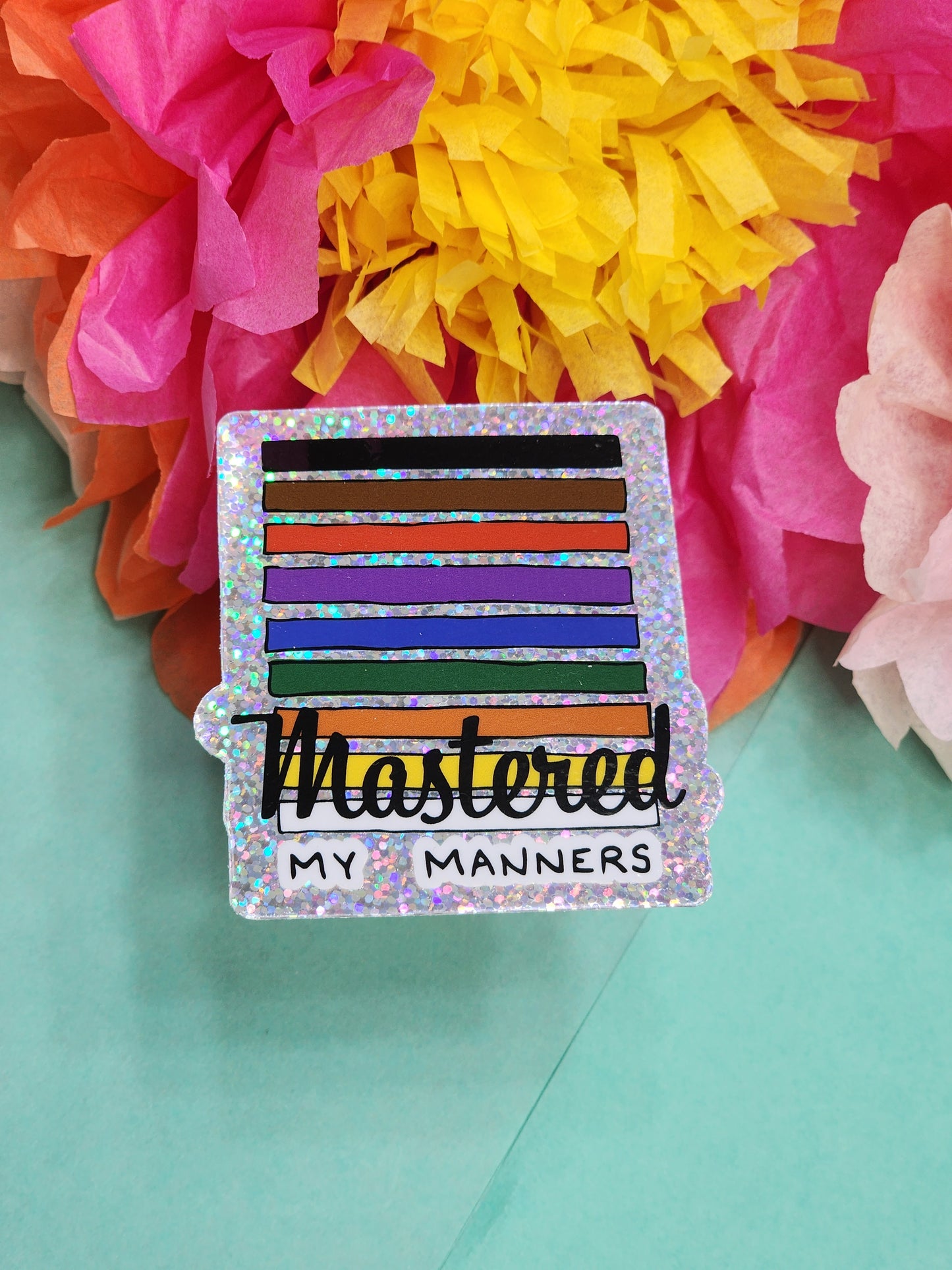 single “Mastered my Manners” glitter sticker on teal surface with bright tissue flowers in the background