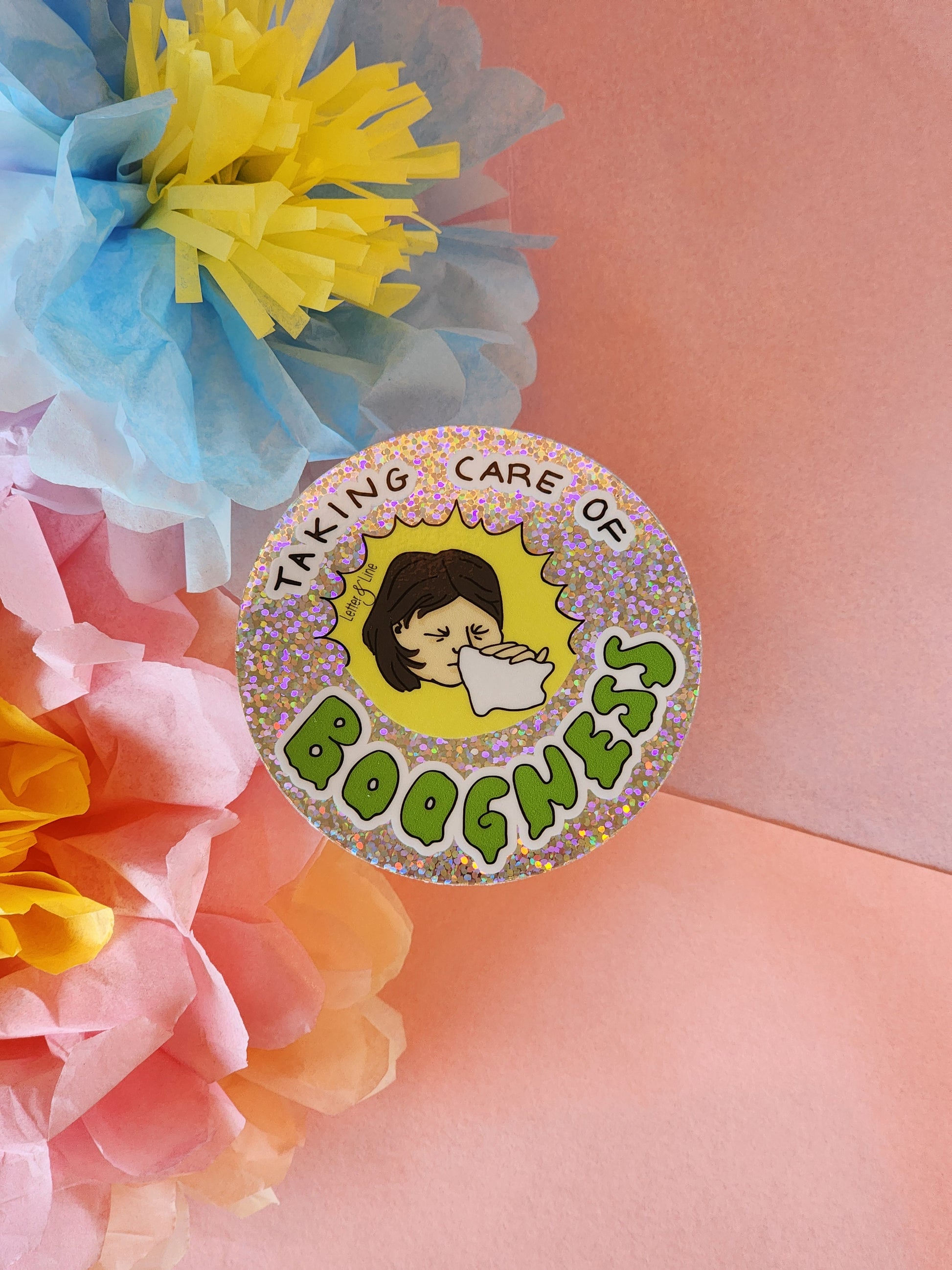 Single “Taking Care of Boogness” round glitter sticker placed on a pink surface with pastel tissue flowers in yellow, blue, and pink.