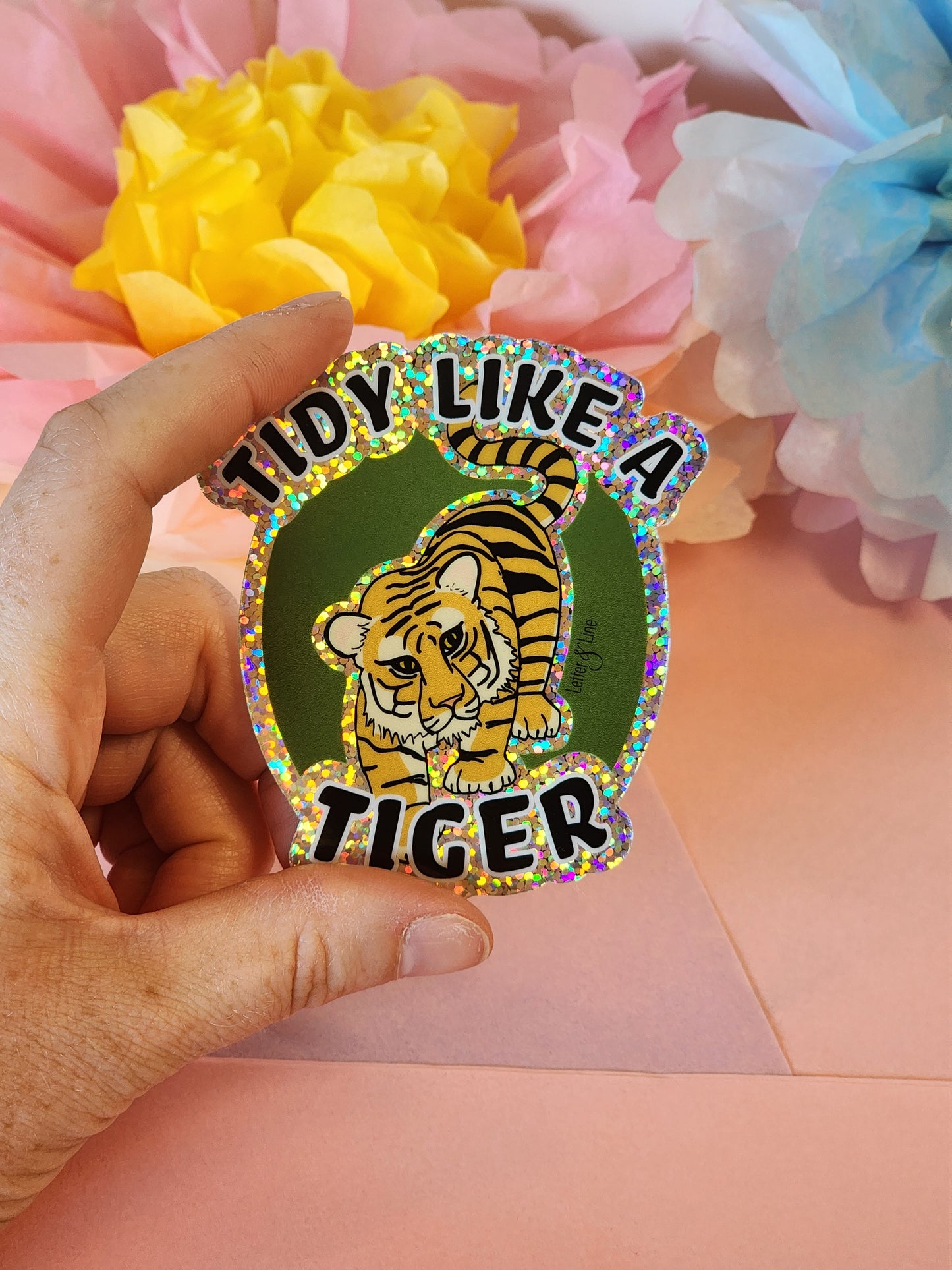 Hand holding a glittery “Tidy Like a Tiger” sticker in front of pastel tissue paper flowers on a pink background.