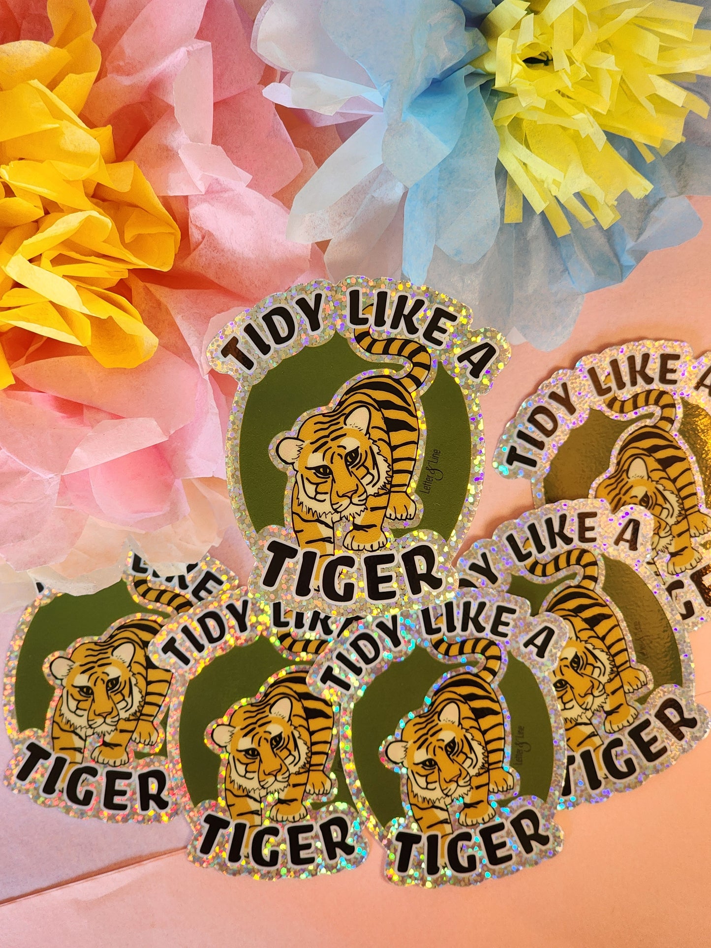 Several “Tidy Like a Tiger” stickers arranged on a pink background with pastel tissue paper flowers in the corners.