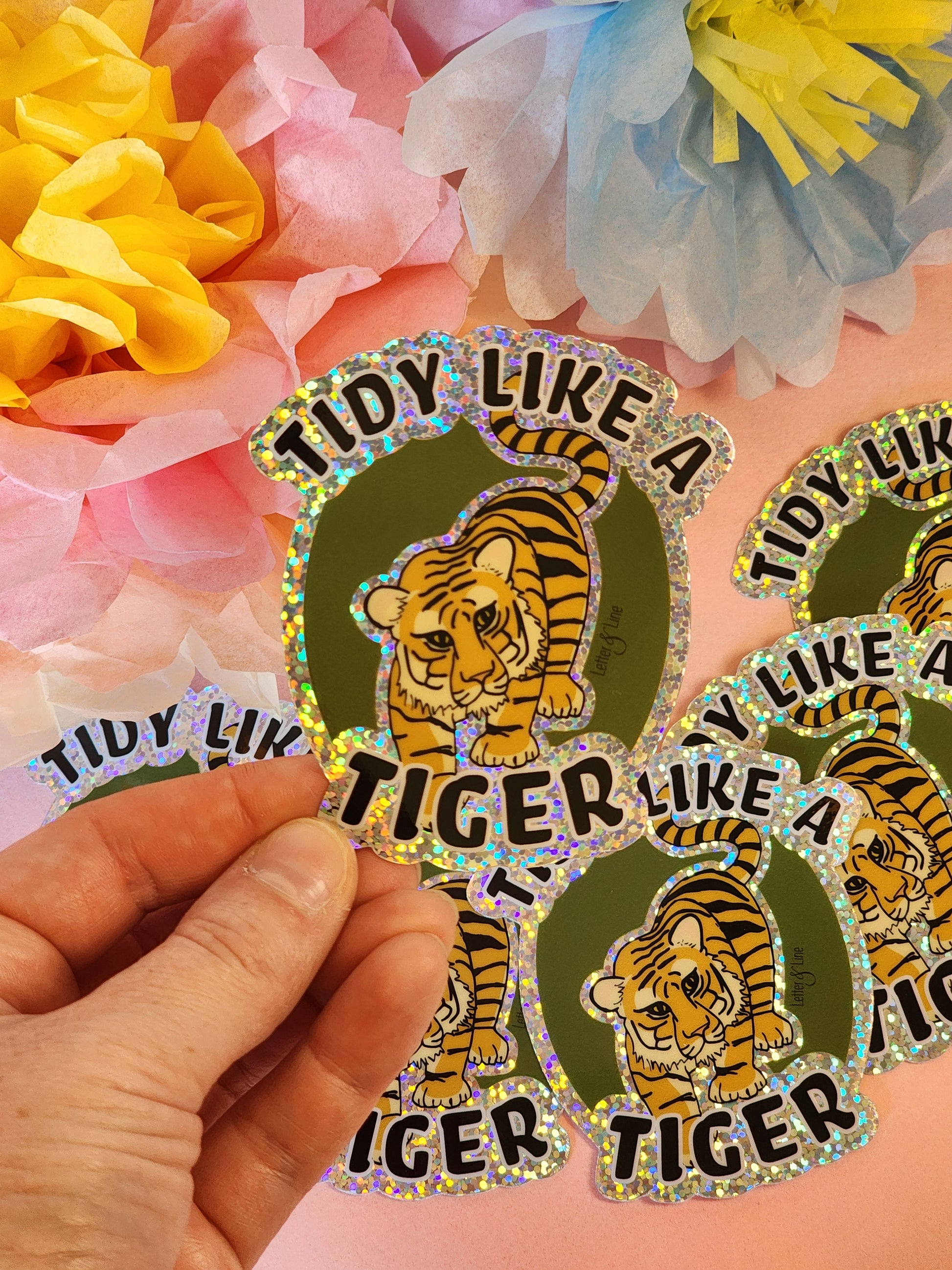 Hand holding one “Tidy Like a Tiger” sticker in front of a scattered group of matching stickers on a colorful background.