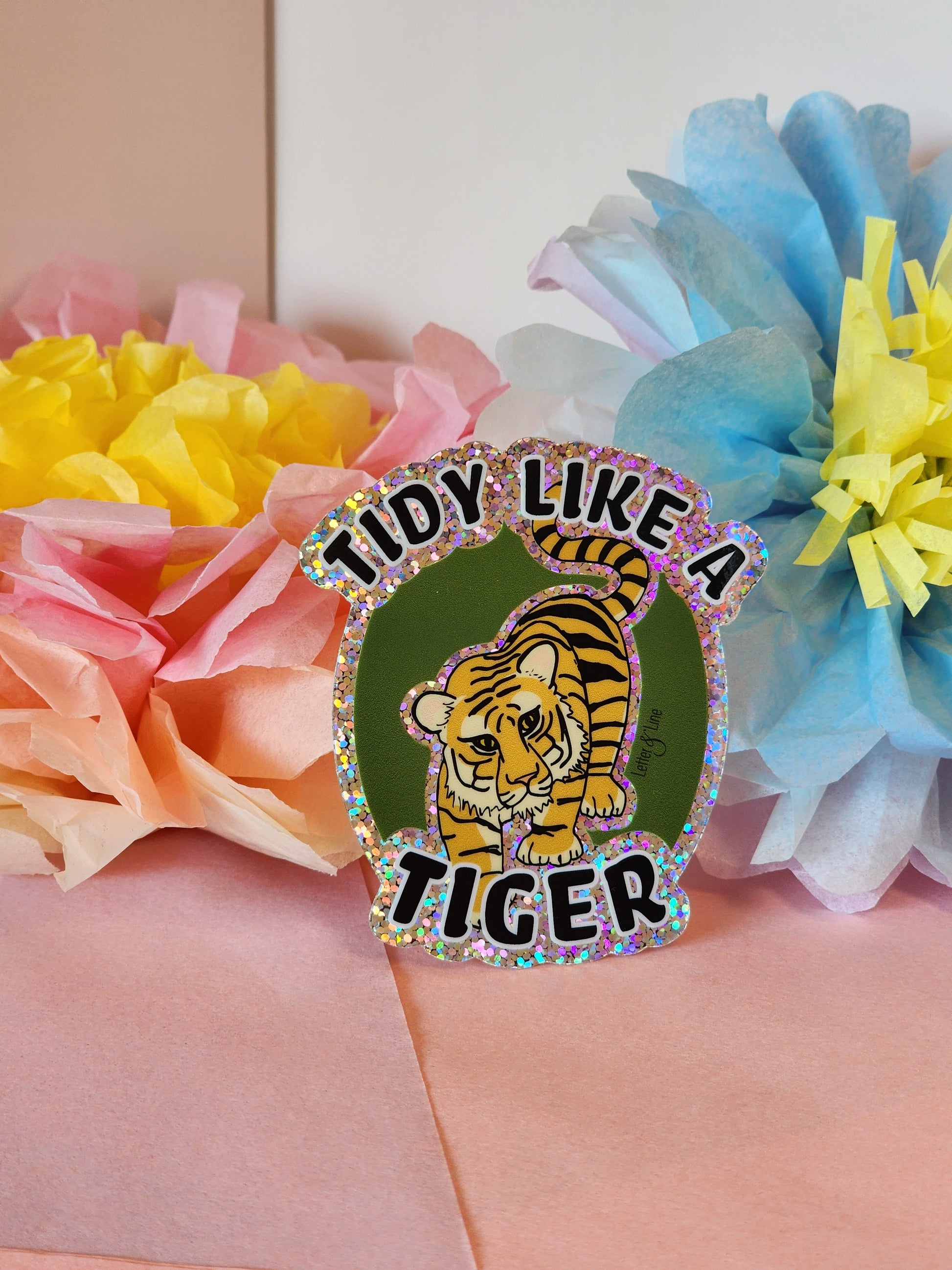 “Tidy Like a Tiger” glitter sticker standing upright in front of soft pastel tissue paper flowers on a light pink surface.