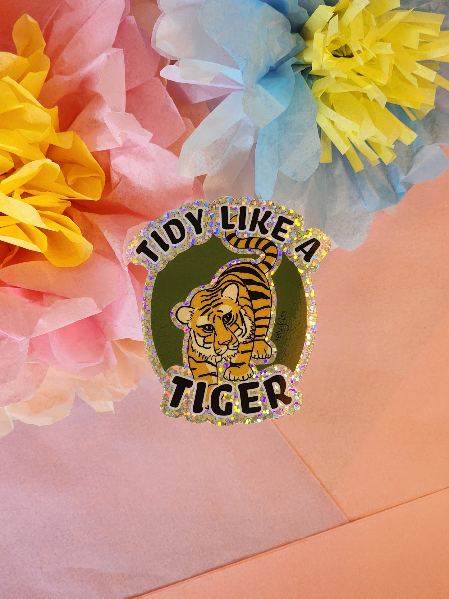 Single “Tidy Like a Tiger” glitter sticker centered on a pink background with yellow and blue tissue paper flowers.